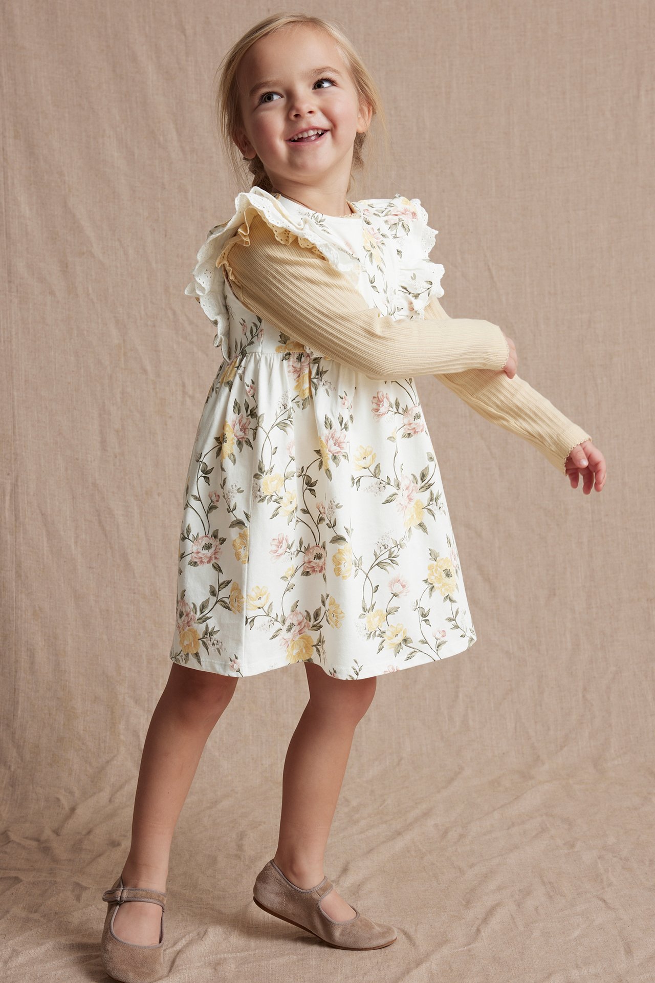 Kind trägt ein weißes geblümtes Kleid mit Rüschen, über einem gelben Rippentop und braunen Schuhen.