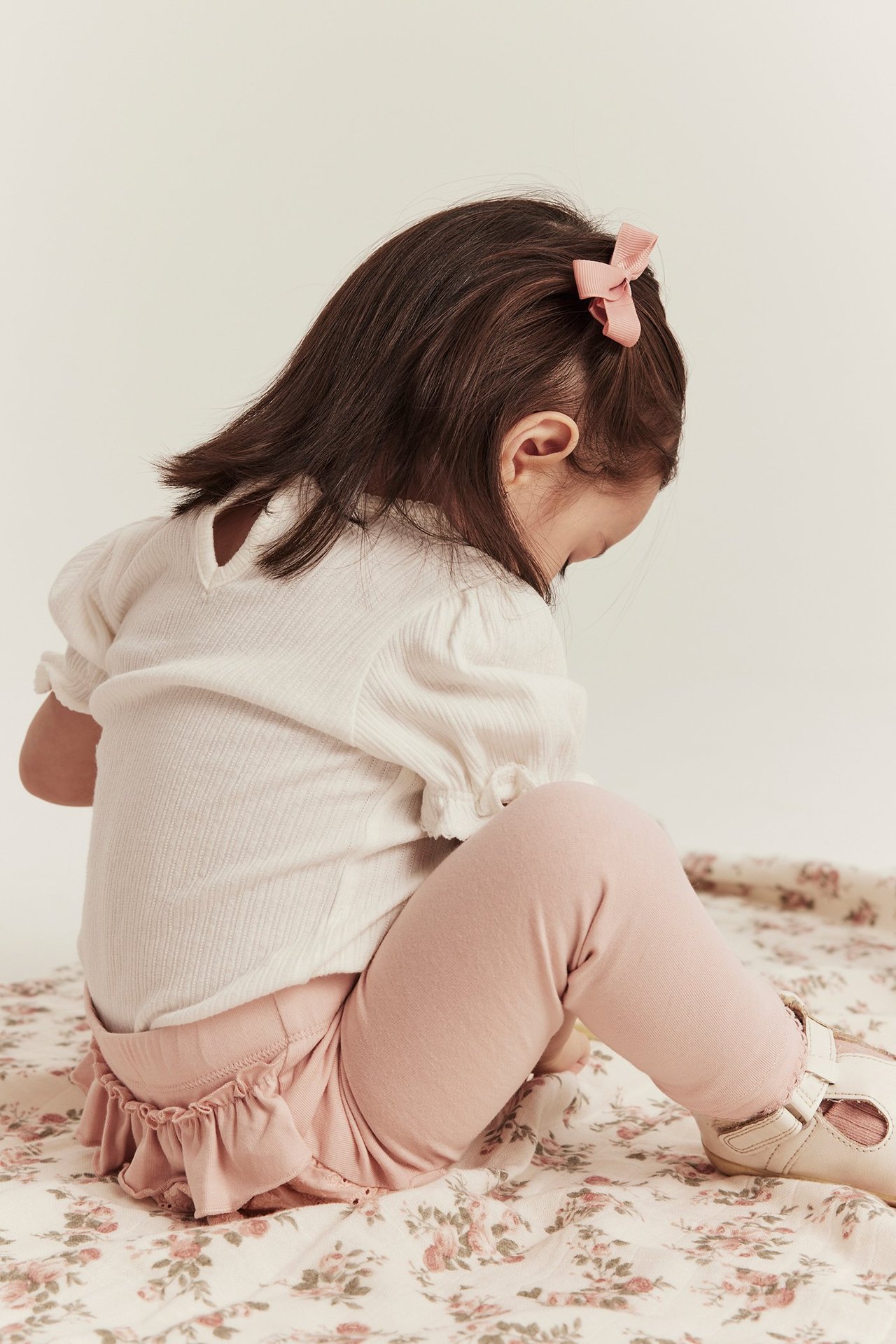 Rückansicht: Kind in hellrosa Rüschenleggings mit Spitze, weißem Rippen-Top, rosa Schleife, hellen Schuhen.