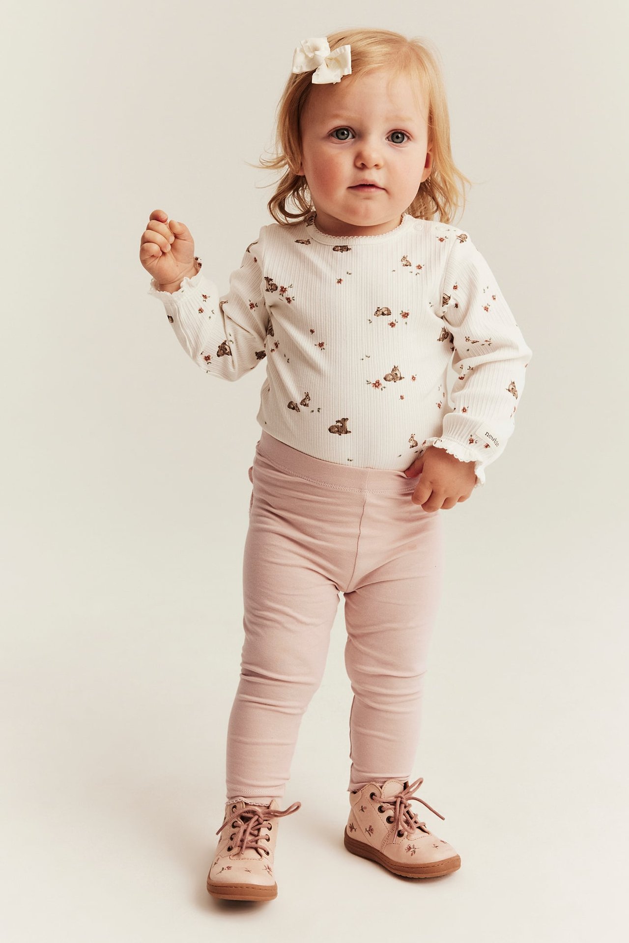 Baby in weißem geripptem Oberteil mit Tiermuster, rosa Leggings und rosa Schnürstiefeln, Vorderansicht.