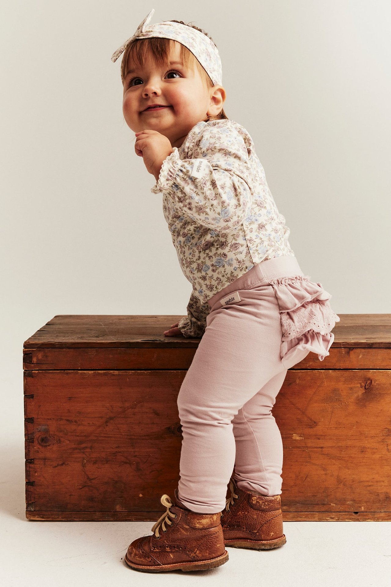 Seitenansicht eines Baby-Mädchens in hellrosa Rüschenleggings, geblümtem Oberteil, passendem Haarband und braunen Stiefeln.