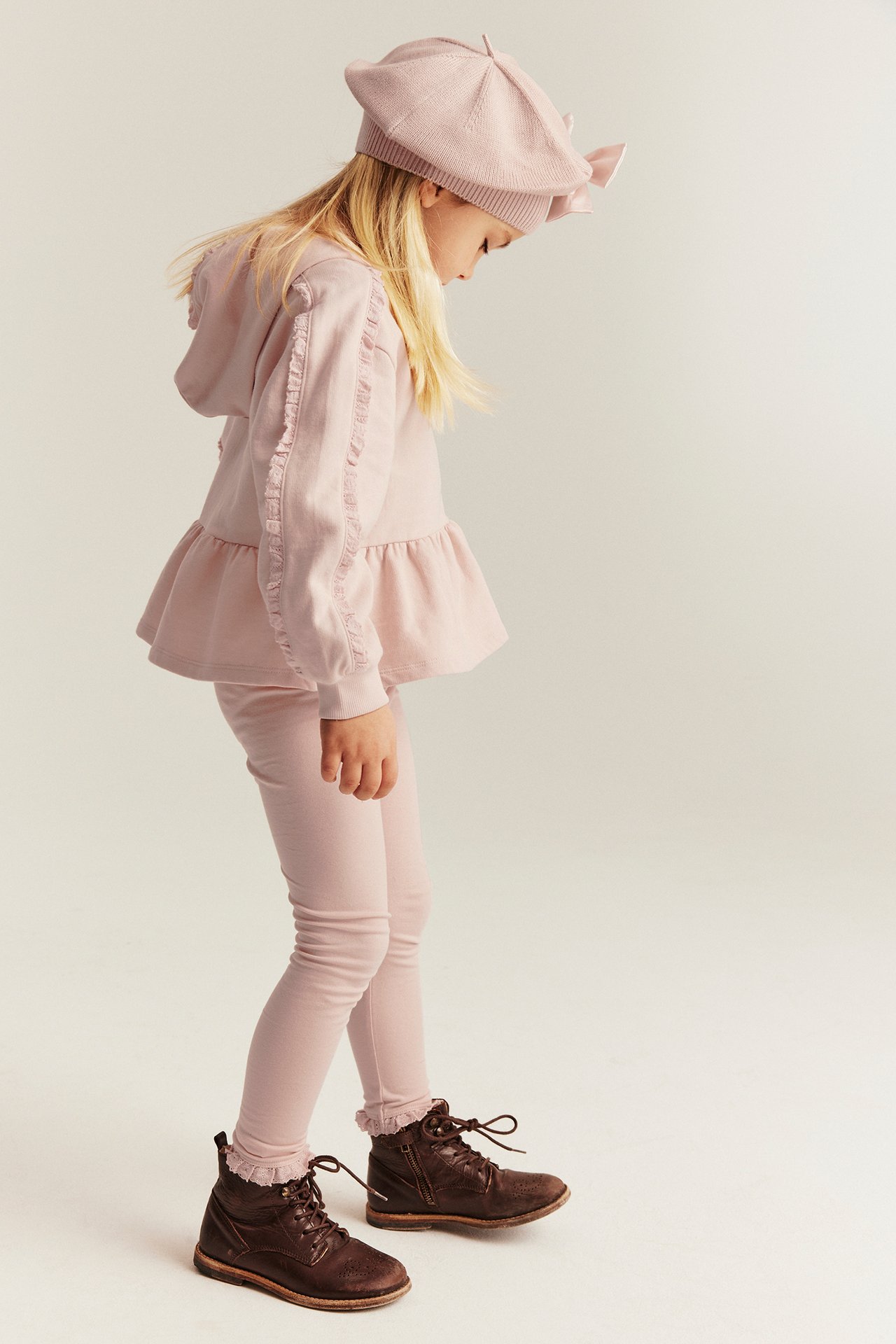 Side view: Kid in light pink leggings with lace, pink ruffled hoodie, beret, and brown boots.