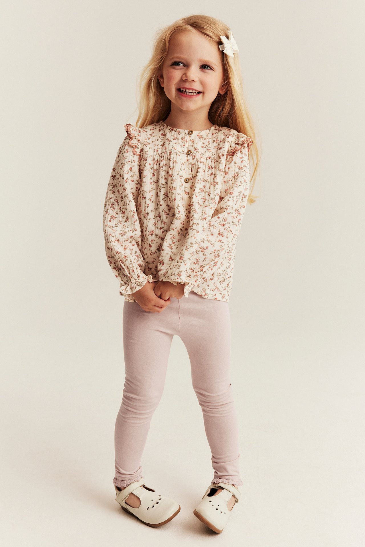 Front view of kid wearing cream floral blouse, pink leggings, and white Mary Jane shoes.