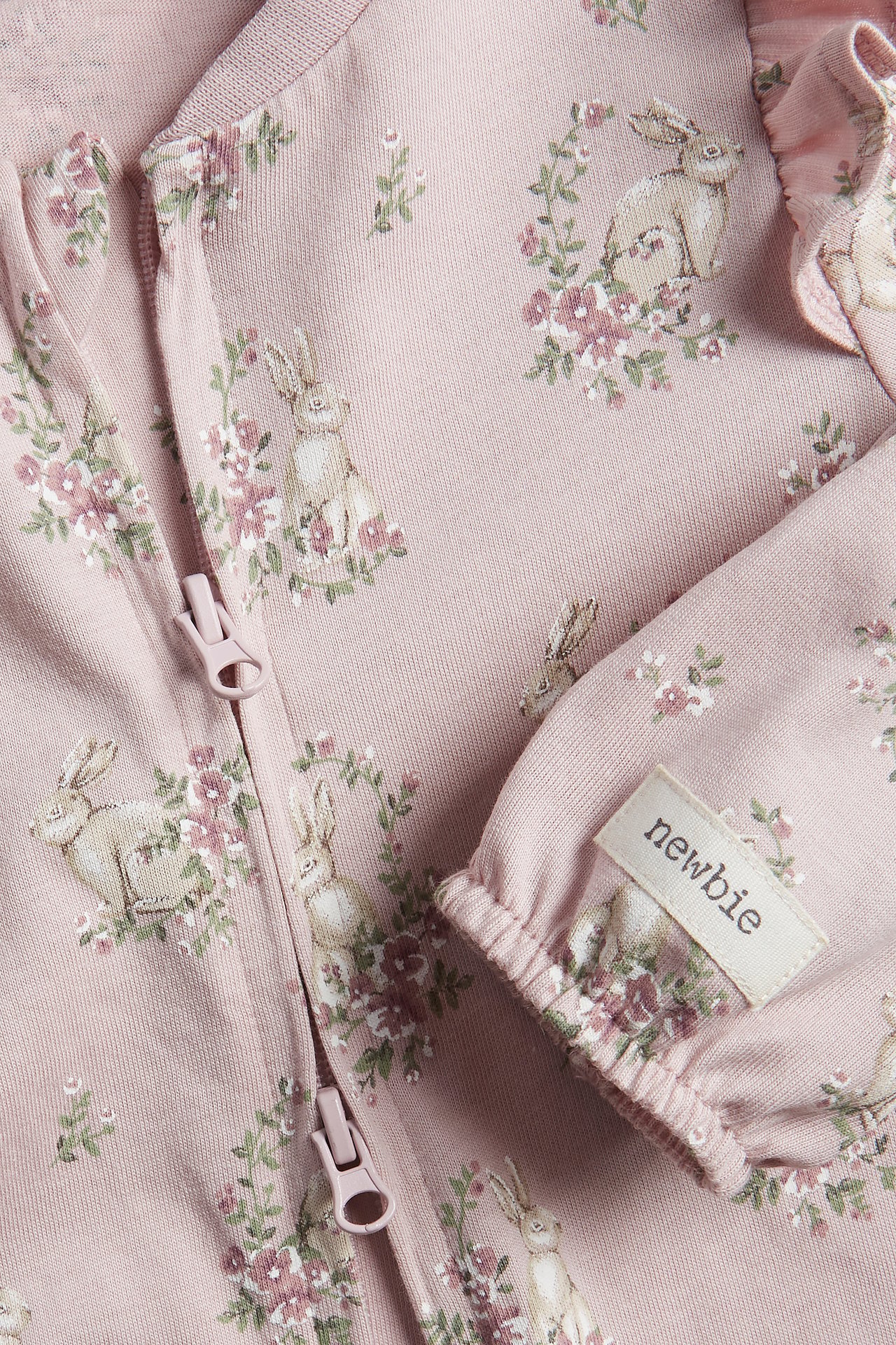Close-up of pink baby pyjamas with rabbit and floral pattern, ruffles on shoulders, and two-way zipper.