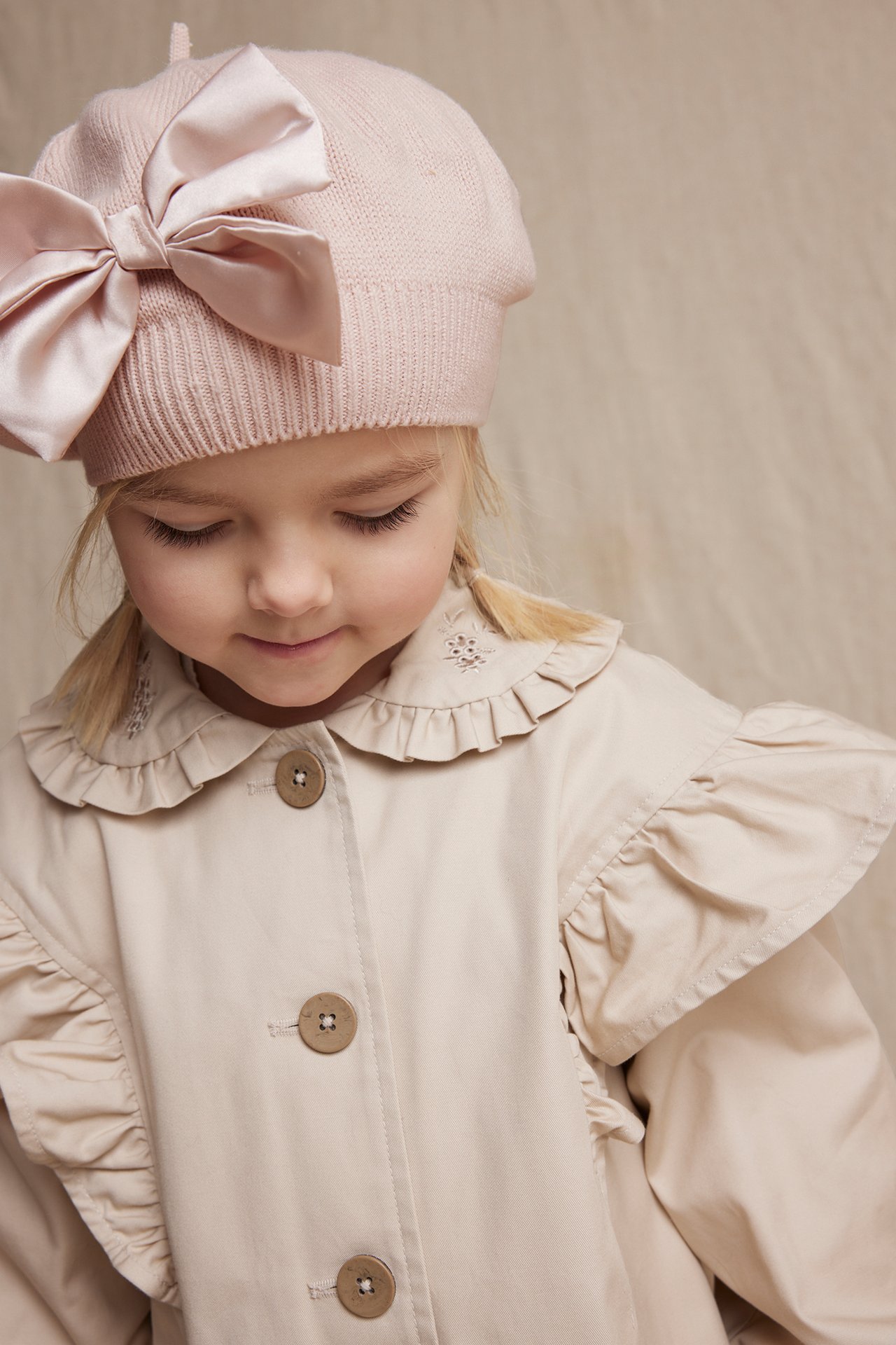 Kid wears a light pink knitted beret with a large shiny bow and a light beige ruffled coat.