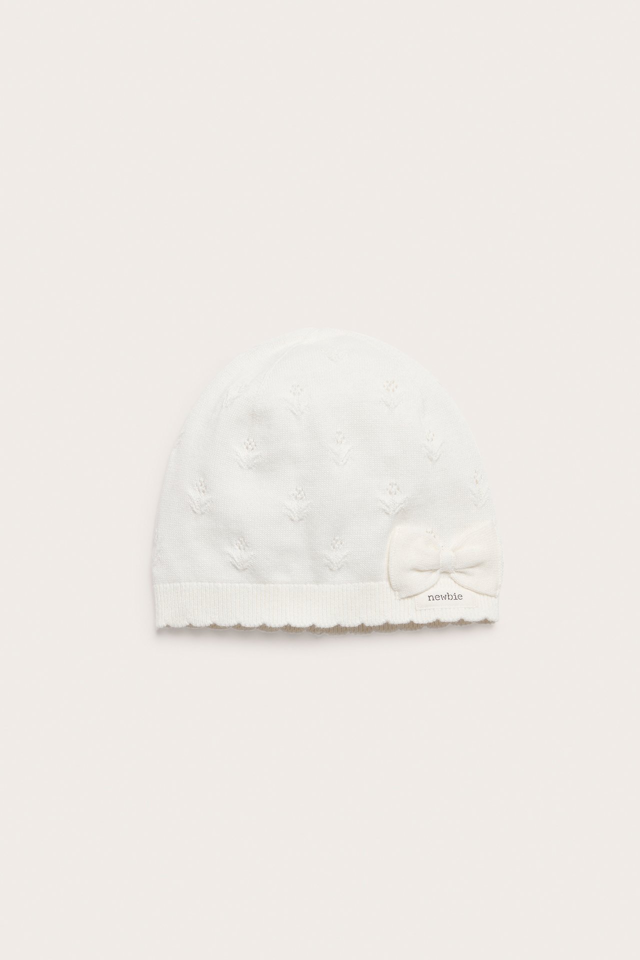 Front view of an off-white baby knitted hat with a subtle pattern, a small bow, and a scalloped edge.