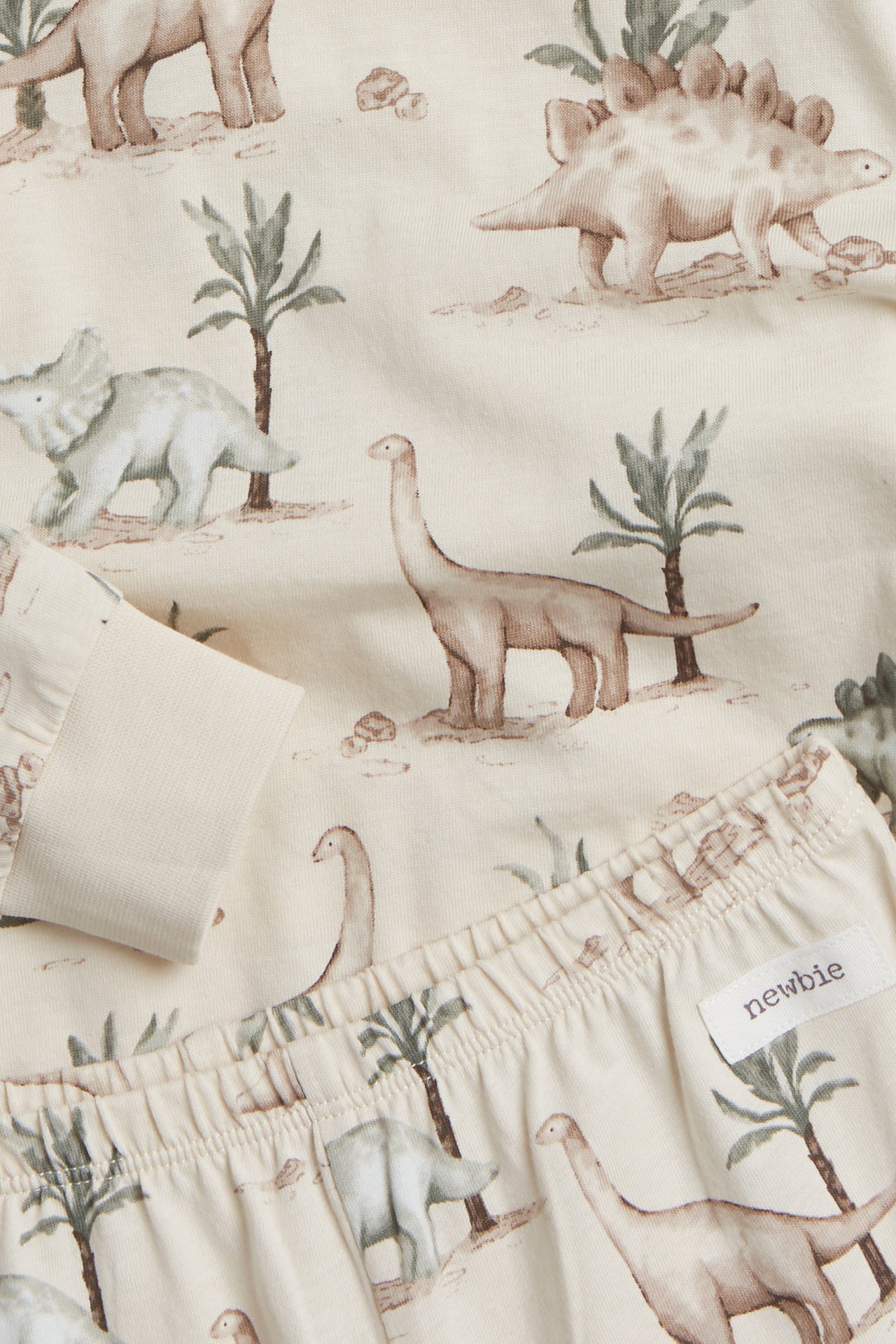 Nahaufnahme eines hellbeigen zweiteiligen Kinder-Jersey-Pyjamas mit Allover-Dinosaurier-Print und Rippbündchen.