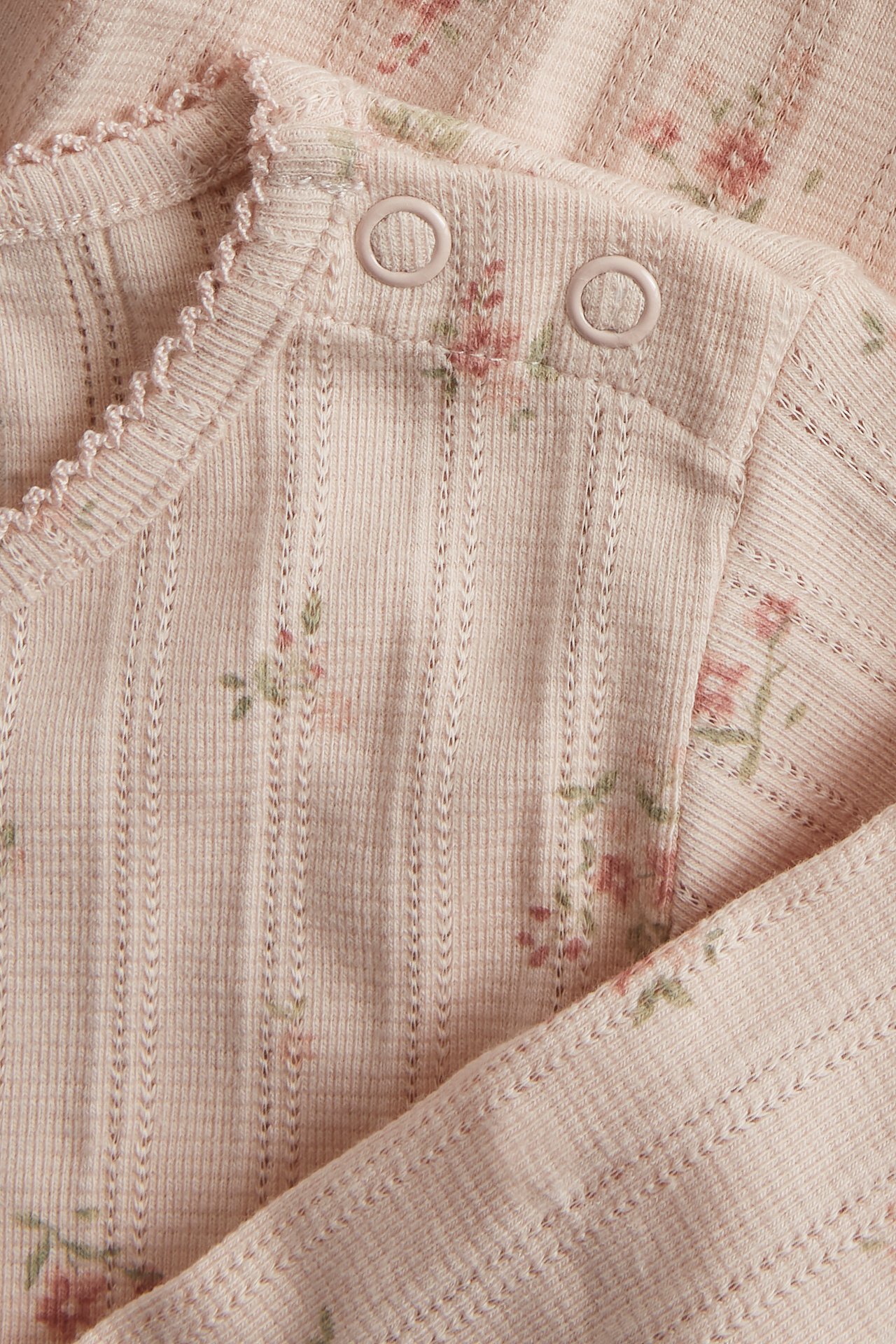 Nahaufnahme eines hellrosa Pointelle-Ripp-Babybodys mit Blumenmuster und Druckknöpfen an der Schulter.