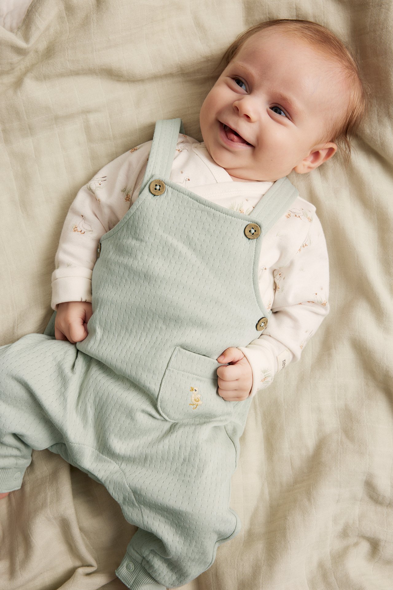 Draufsicht auf ein lächelndes Baby in hellgrüner strukturierter Jersey-Latzhose mit Stickerei über einem Body.