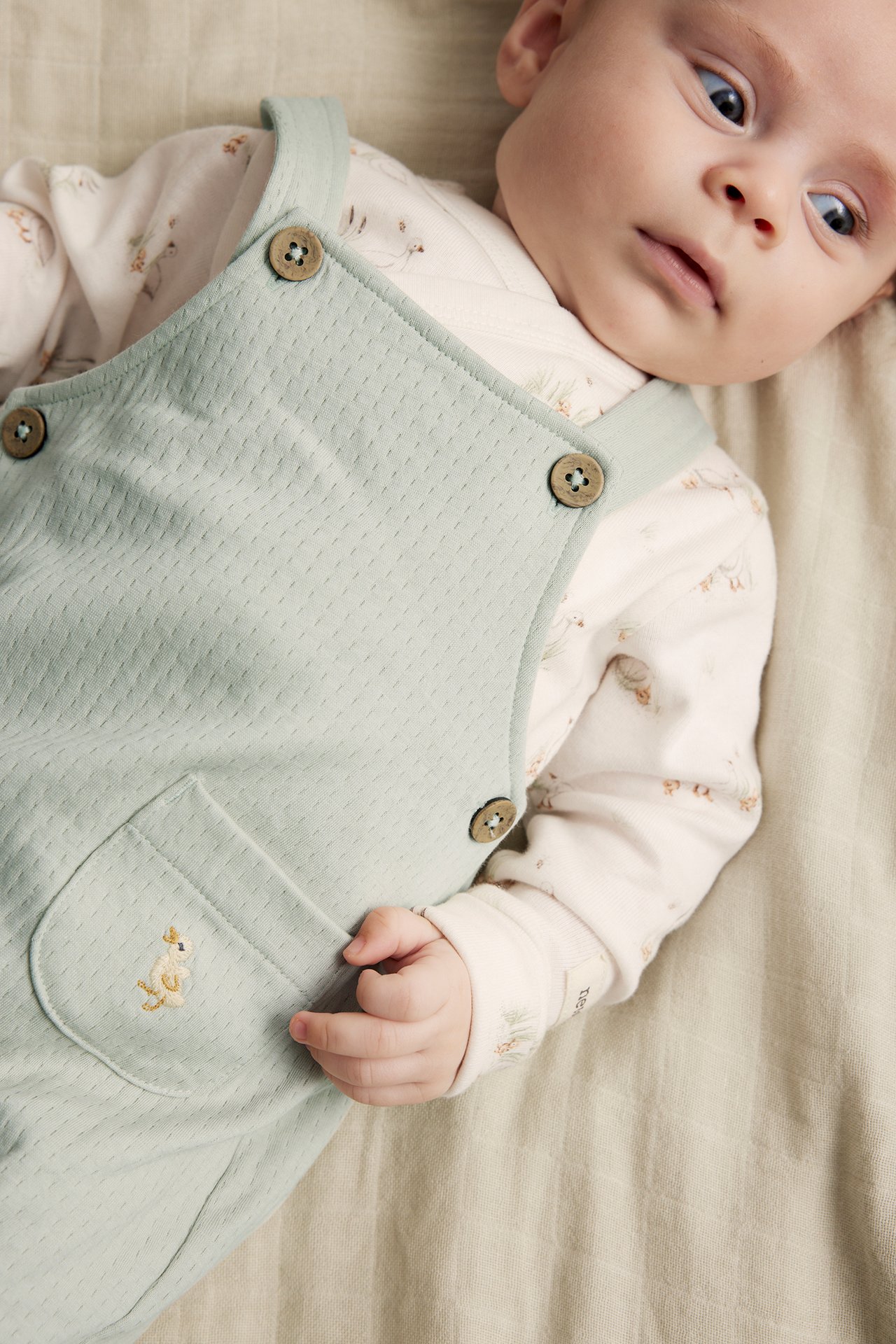 Nahaufnahme eines Babys in hellgrünen Jersey-Latzhosen mit Entenstickerei über einem weißen gemusterten Langarm-Body.