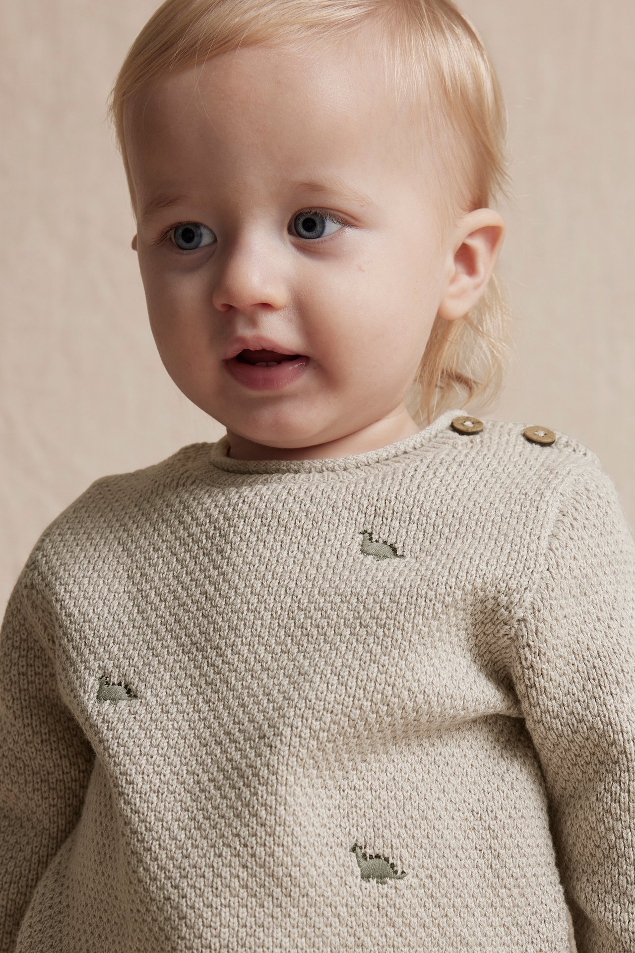 Close-up front view of a baby wearing a beige knitted cotton sweater with dinosaur embroidery and shoulder buttons.