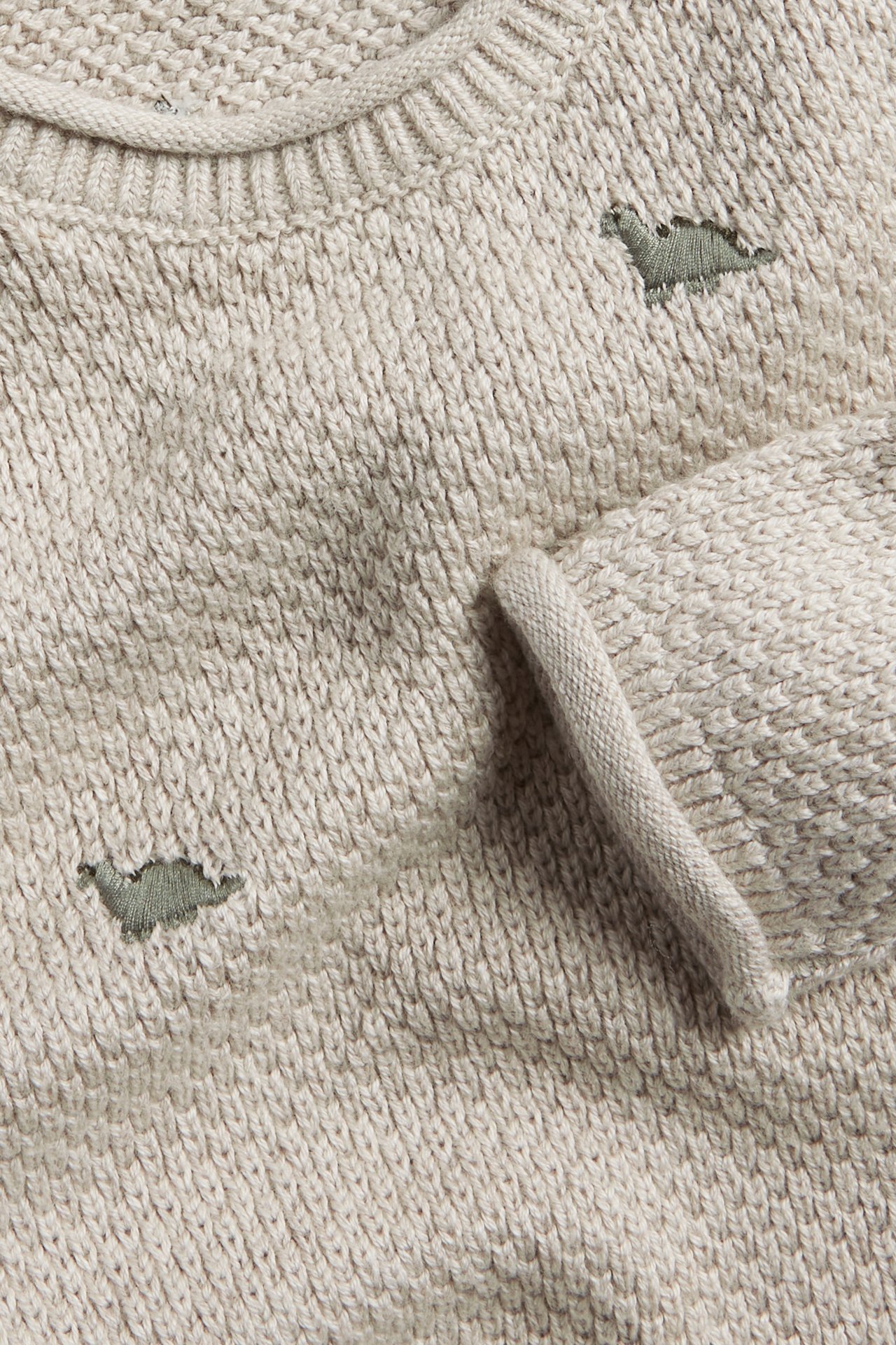 Close-up of a beige melange knitted cotton sweater with green dinosaur embroidery and ribbed details.