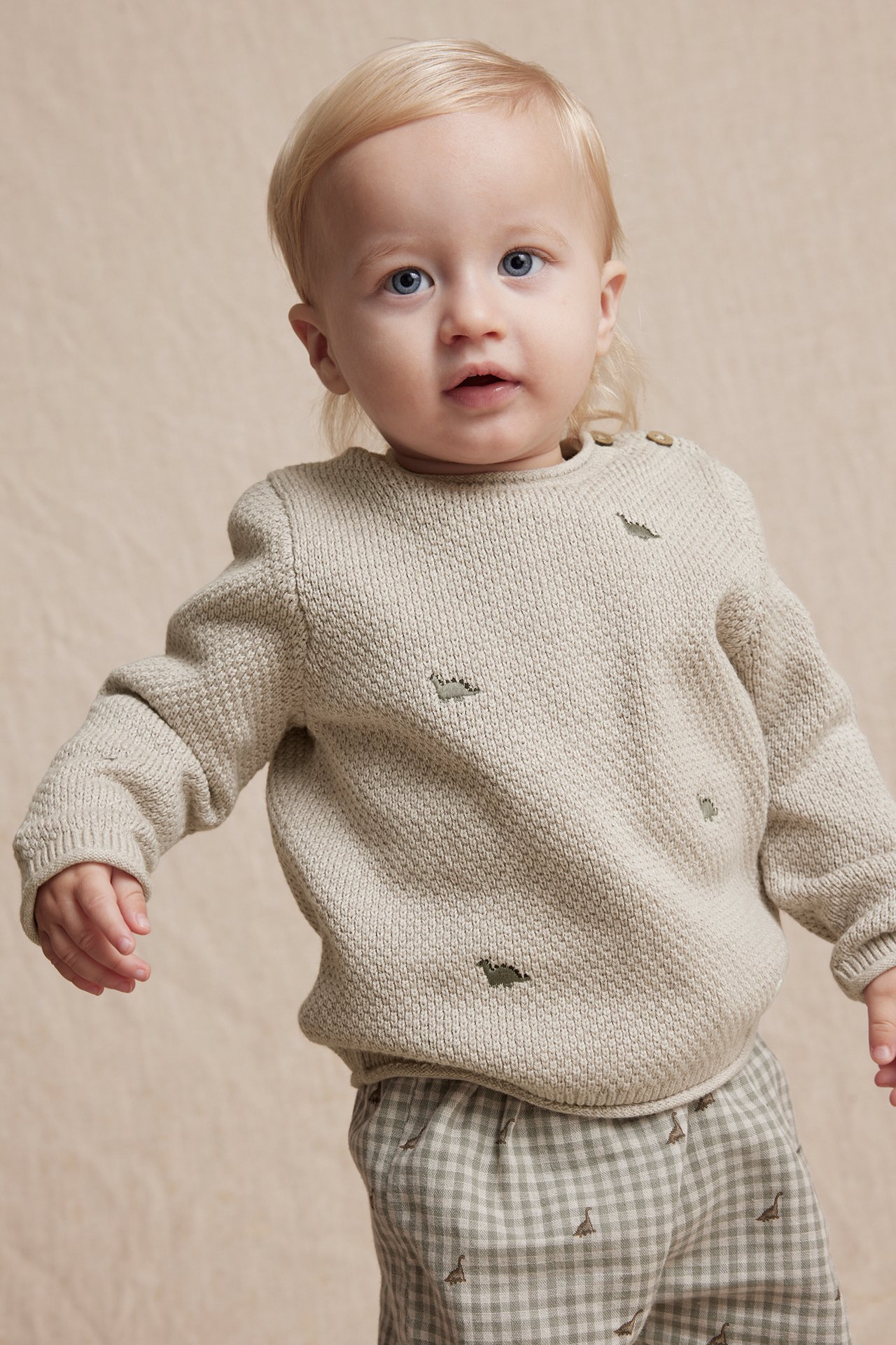 Front view of a baby wearing a beige knitted cotton sweater with dinosaur embroidery and checkered pants.