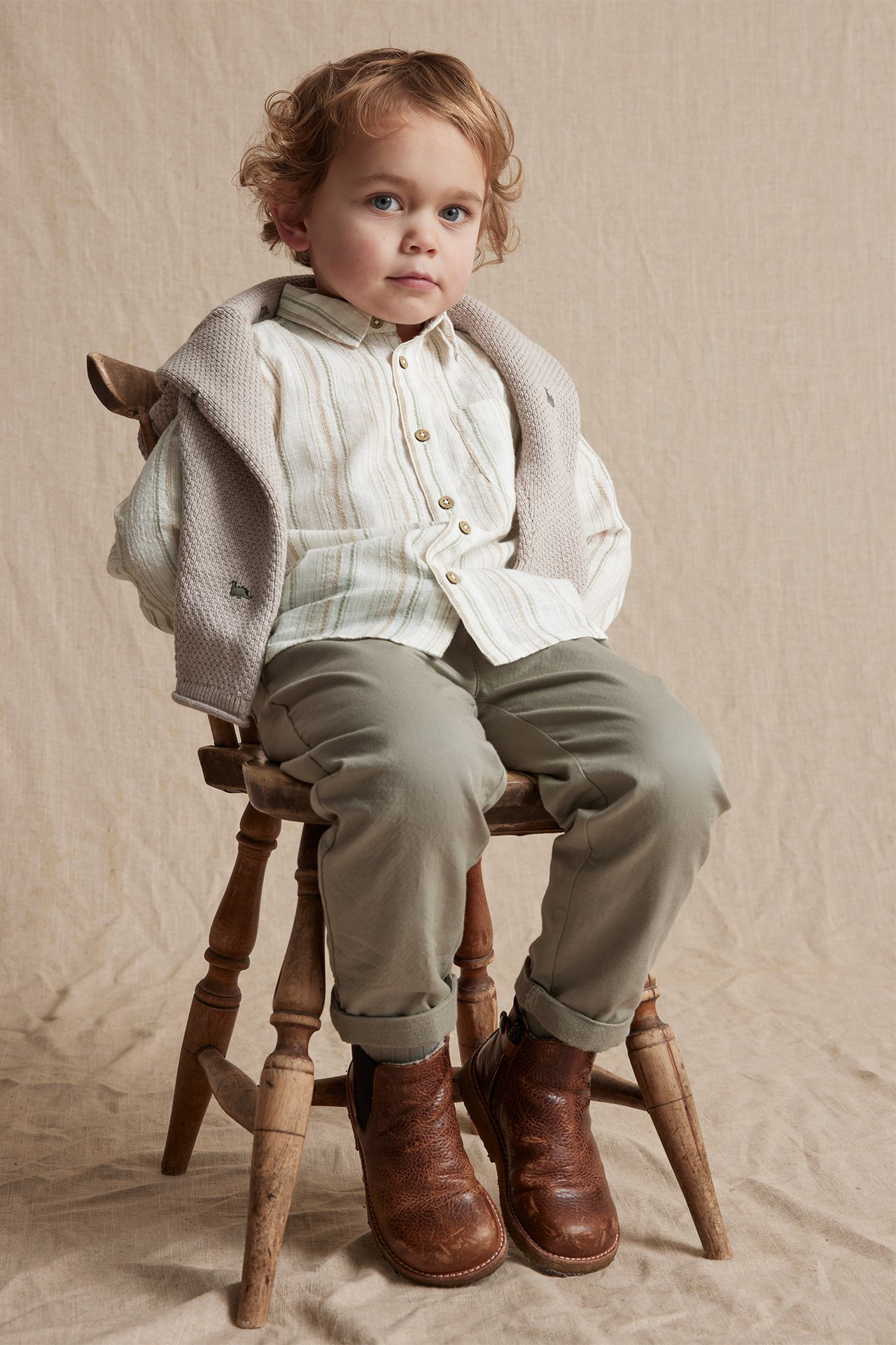 Vorderansicht: Kind in cremeweißem gestreiftem Baumwollhemd, hellgrüner Hose, braunen Stiefeln und grauem Strickcardigan.