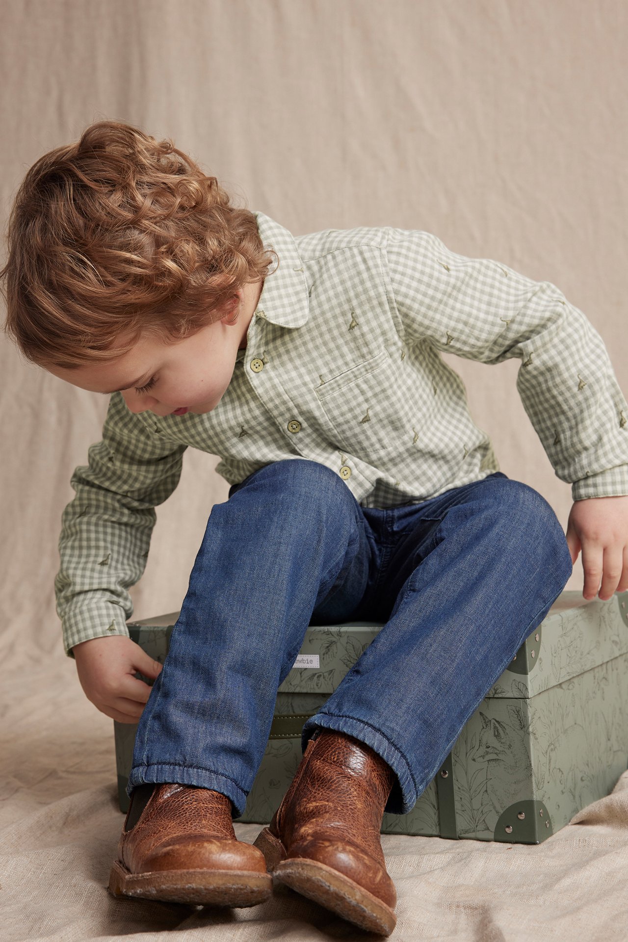 Kid in light green and white checked cotton shirt with embroidery, pocket, blue jeans, and brown boots.