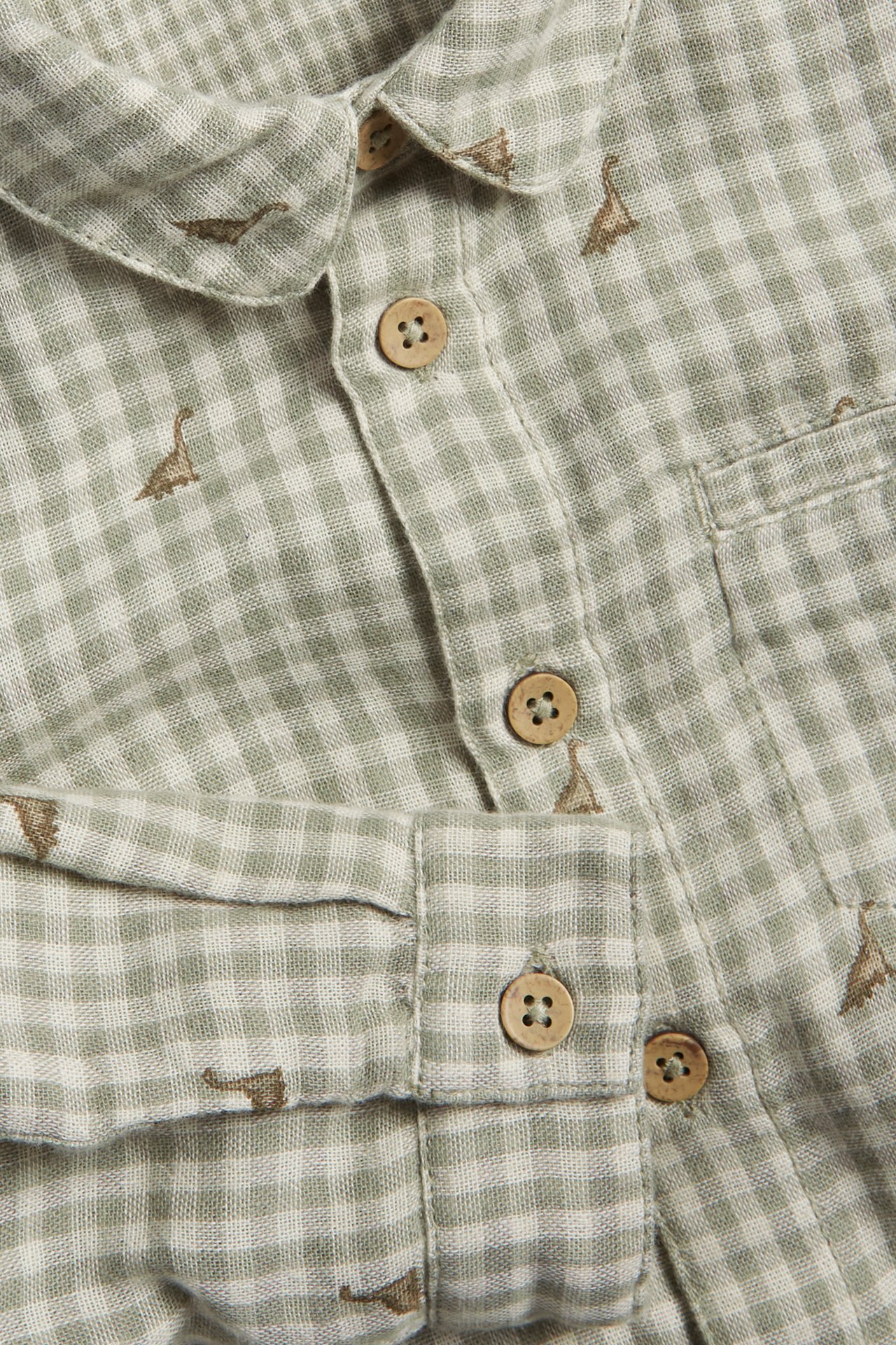 Close-up of a light green and white checked cotton shirt with brown dinosaur embroidery, front buttons, and a pocket.