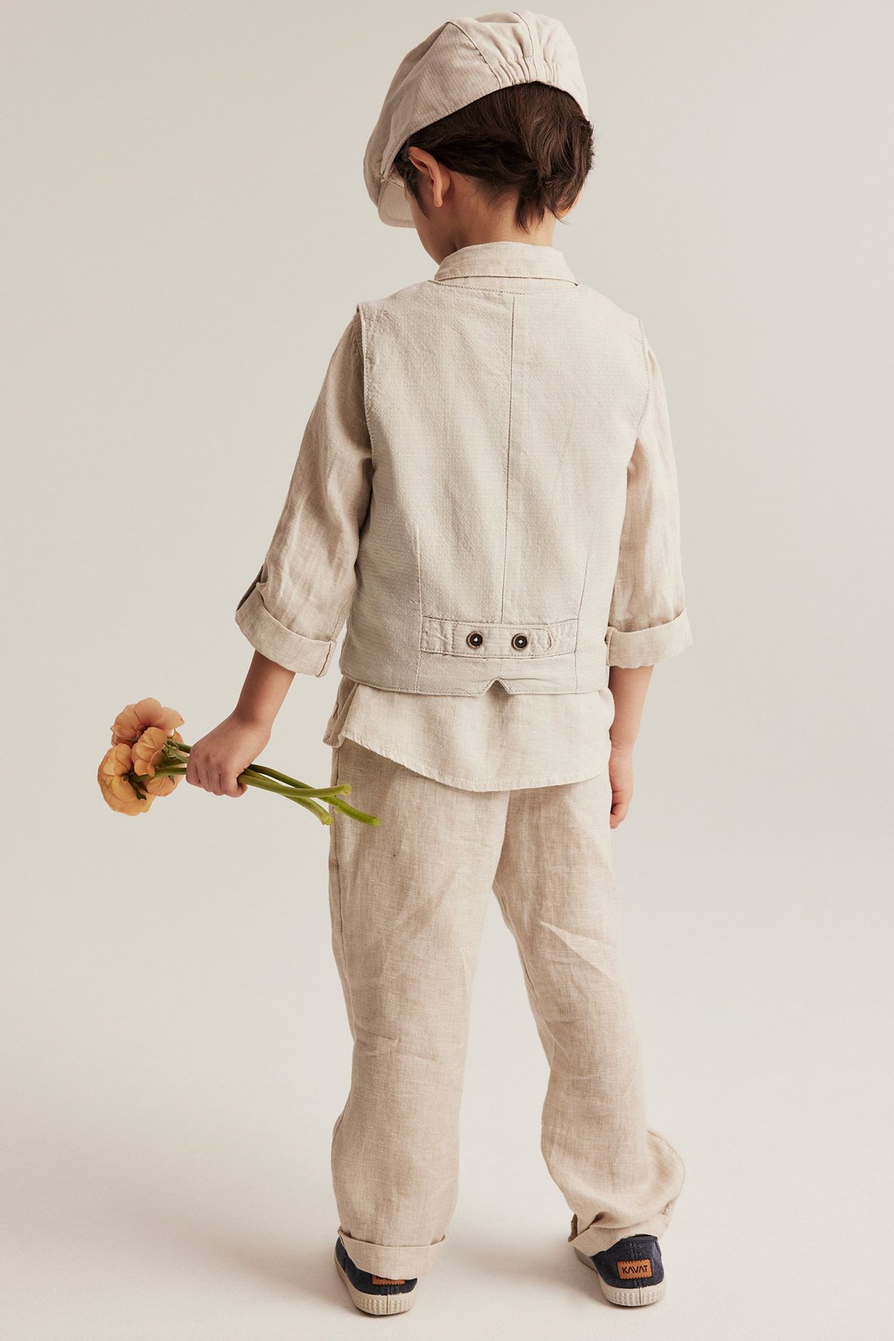 Rückenansicht Kind: beige Webweste mit Rückenspange, Hemd, Hose, Mütze; hält orange Blumen.