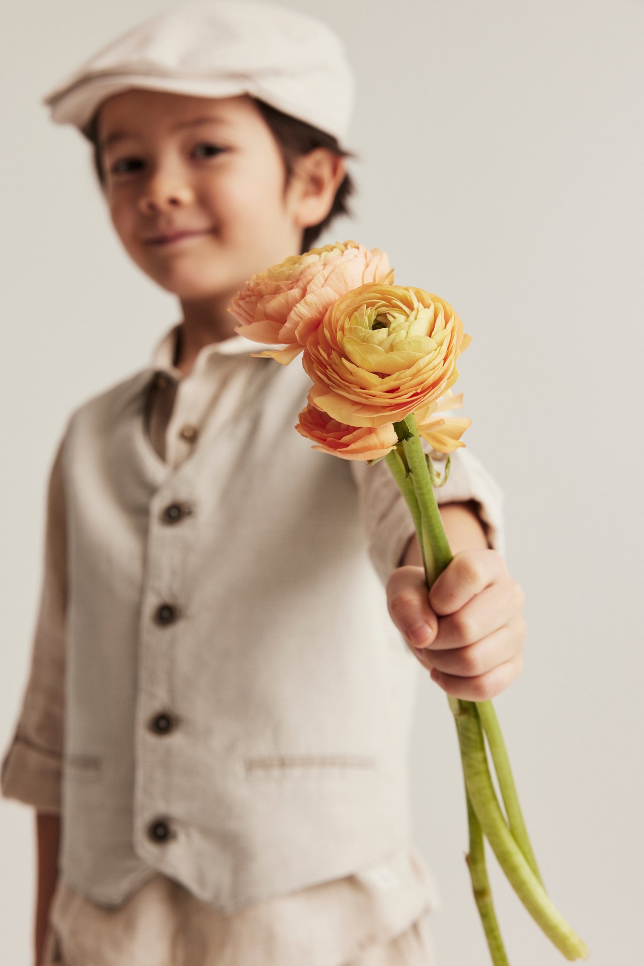 Vorderansicht: Kind in beiger gewebter Weste mit Knöpfen, beigem Hemd, Mütze, hält orange Blumen.