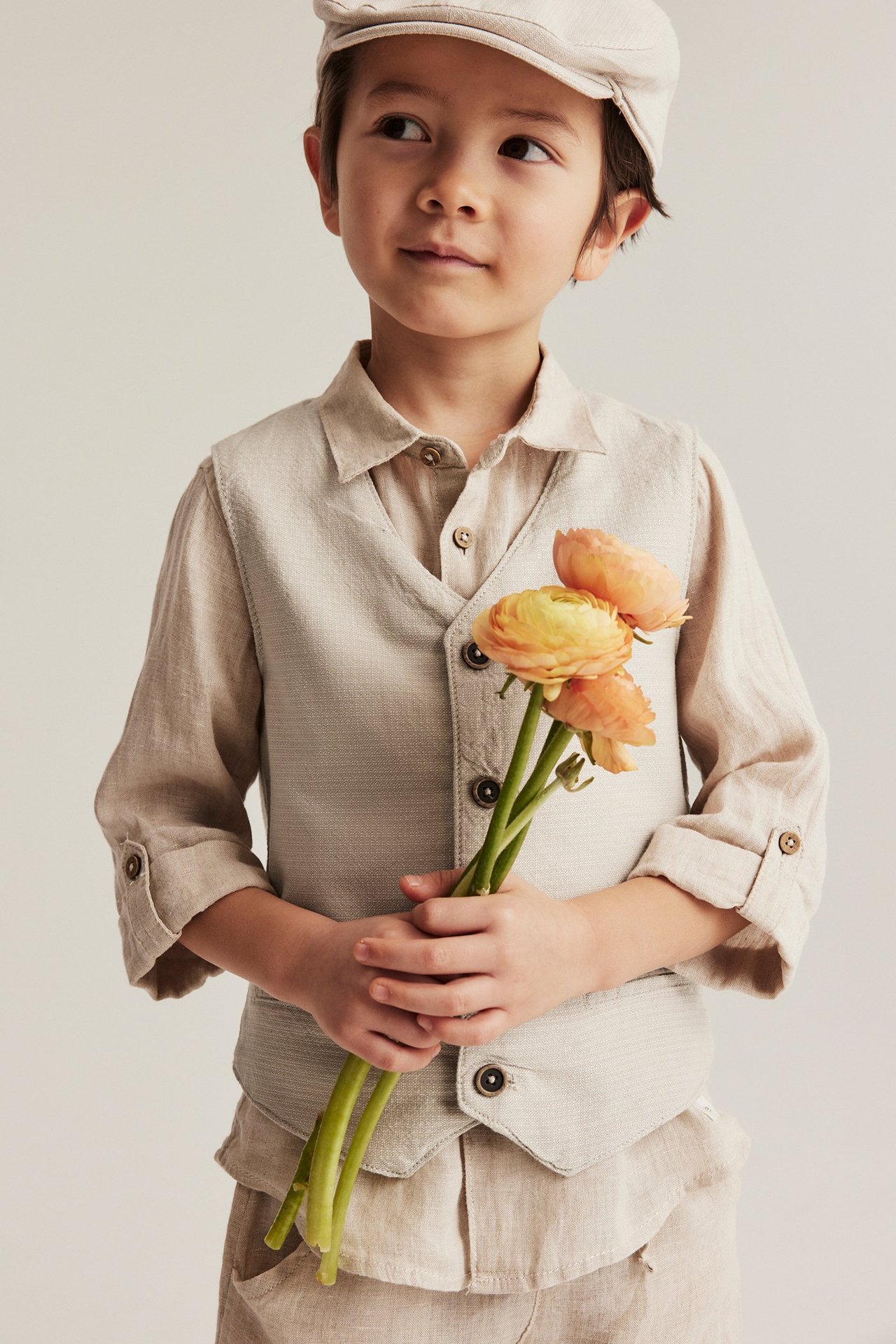 Kind in beiger gewebter Weste, passendem Hemd, Hose und Mütze, hält orangefarbene Blumen.