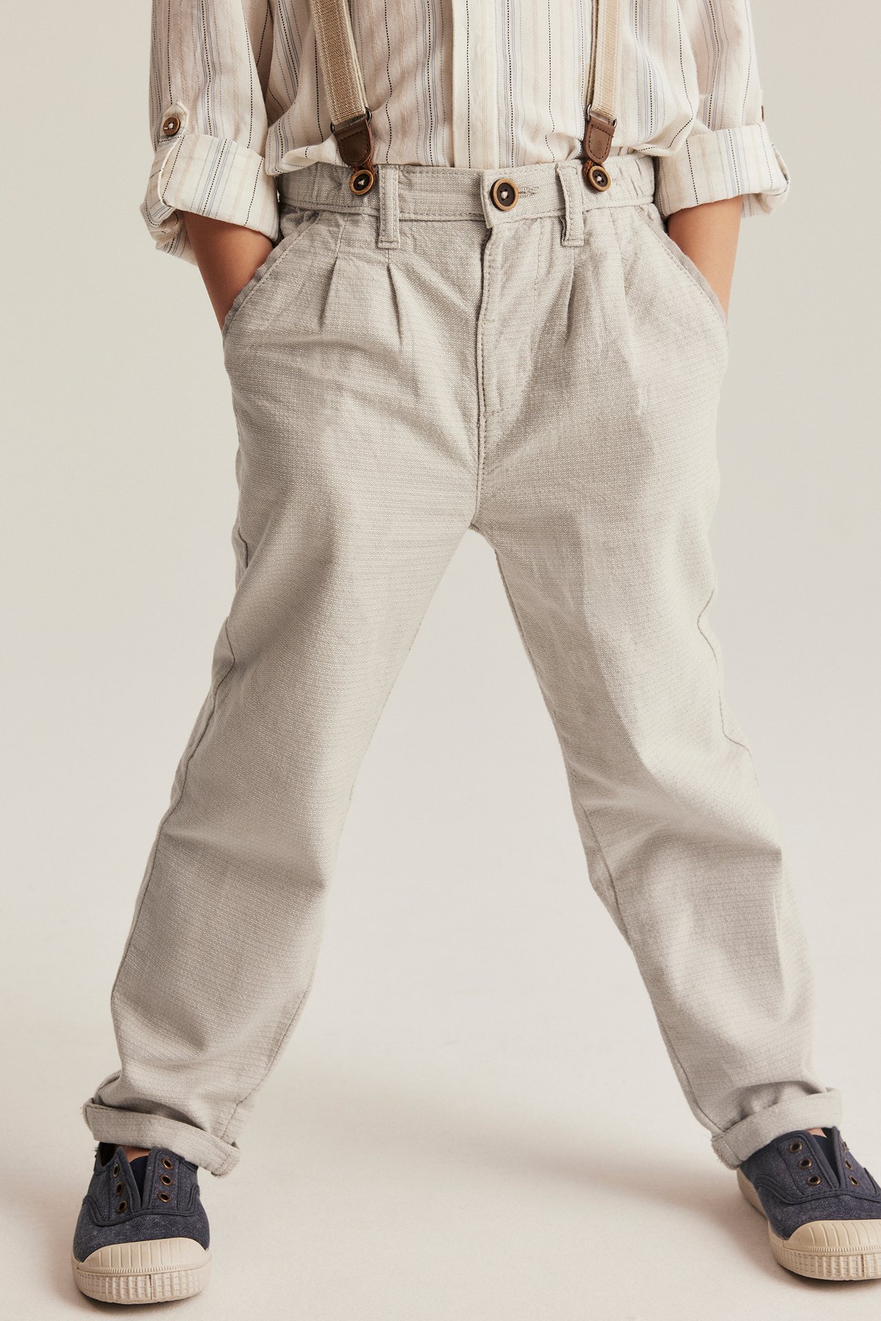 Front view: Kid in beige woven trousers with brown suspenders, striped shirt, and dark blue shoes.