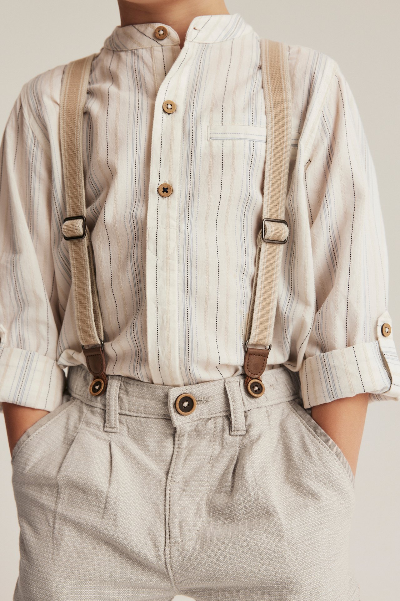 Front view of a kid wearing beige woven trousers with suspenders over a striped shirt.