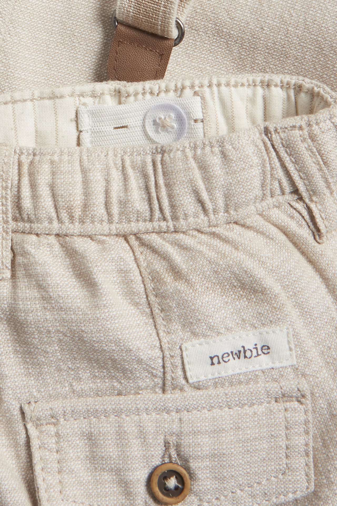 Close-up of beige woven cotton dungaree trousers with elasticated waist, decorative button, and suspenders.