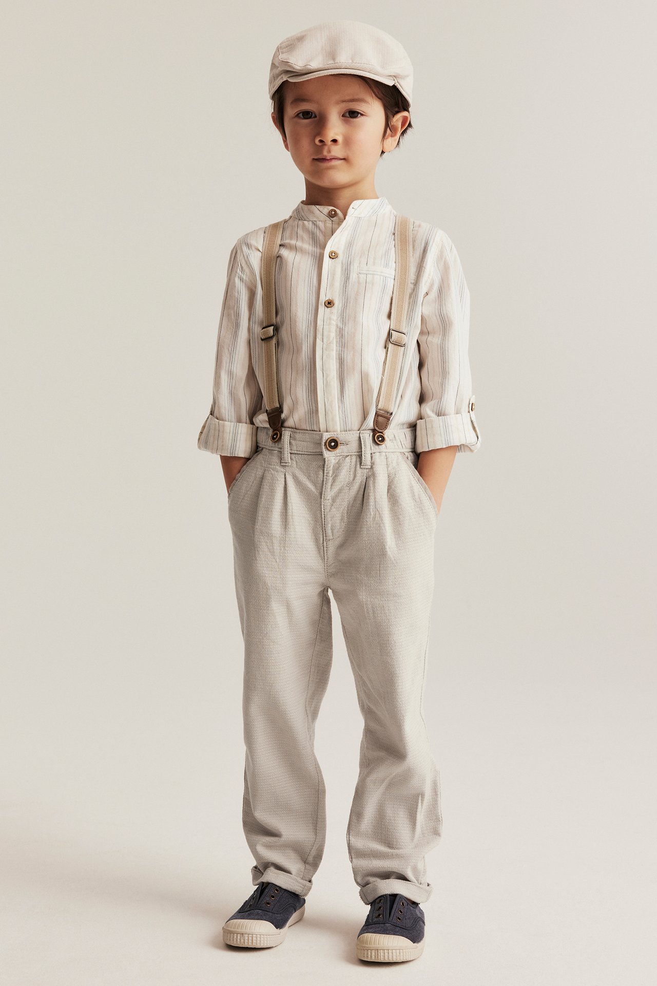Front view: Kid in beige woven trousers with suspenders, striped shirt, beige cap, and dark blue shoes.