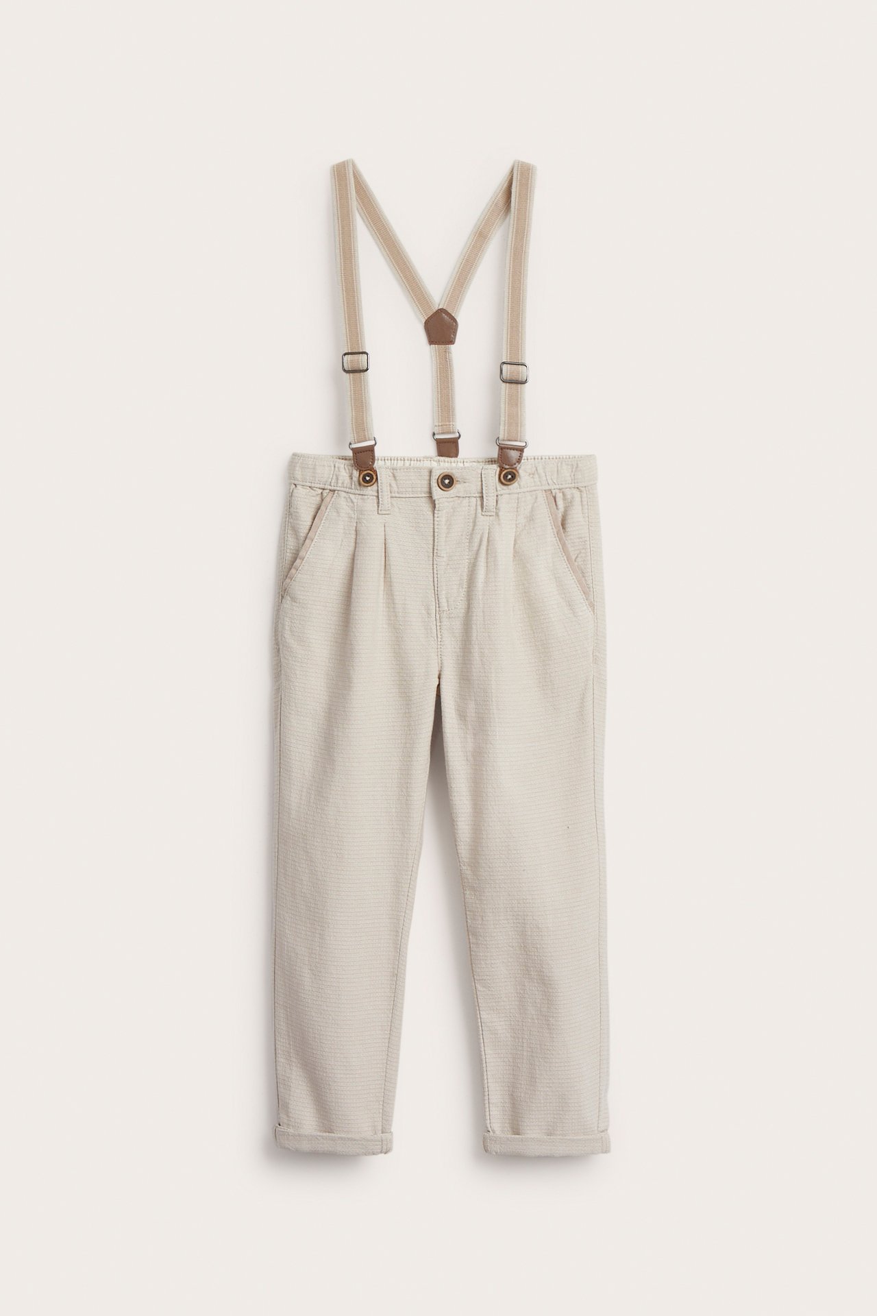 Front view of kid's beige woven trousers with adjustable suspenders and rolled-up cuffs.