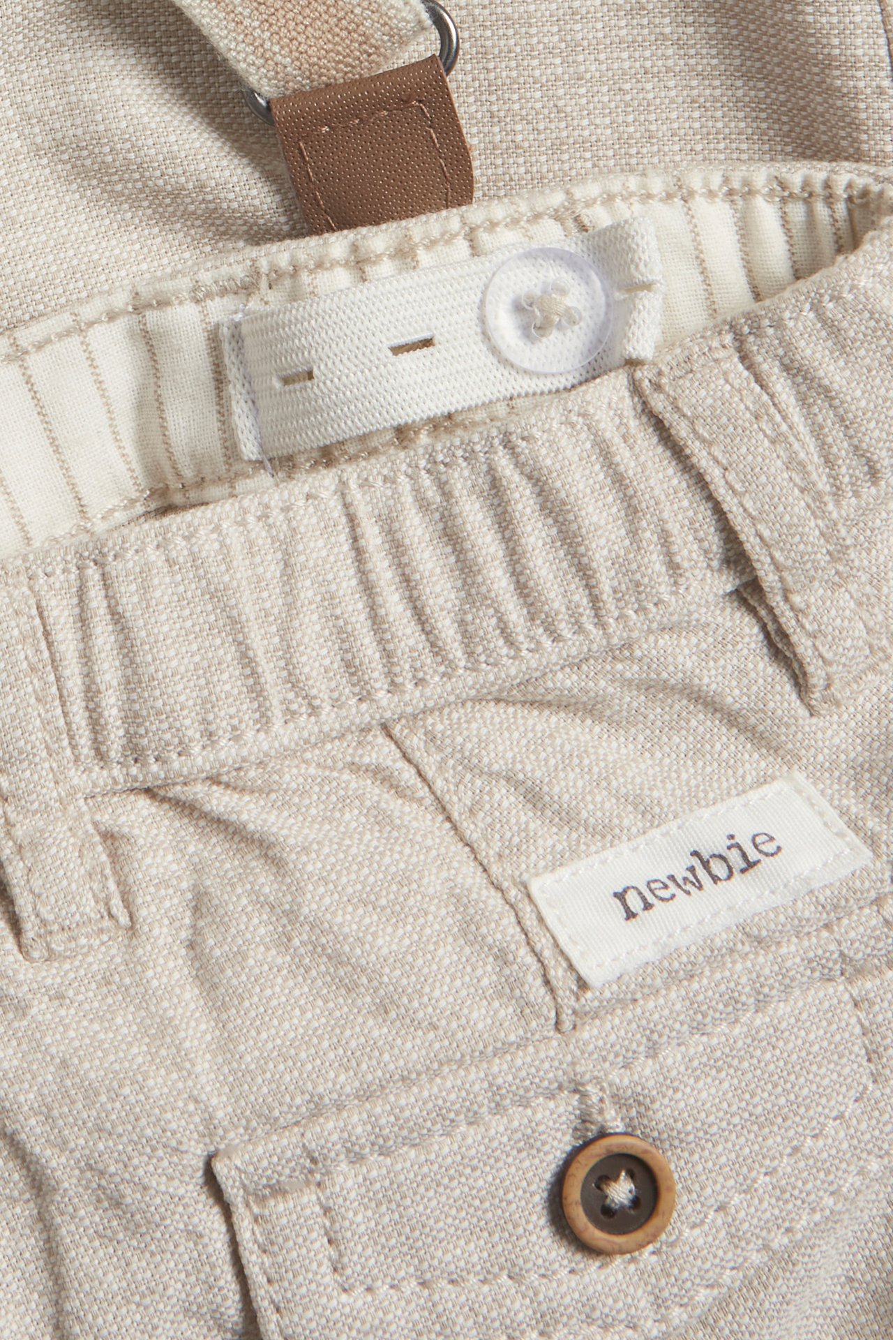 Nahaufnahme beiger gewebter Kinderhose mit verstellbarem Bund, transparentem Knopf und braunem Hosenträgerdetail.