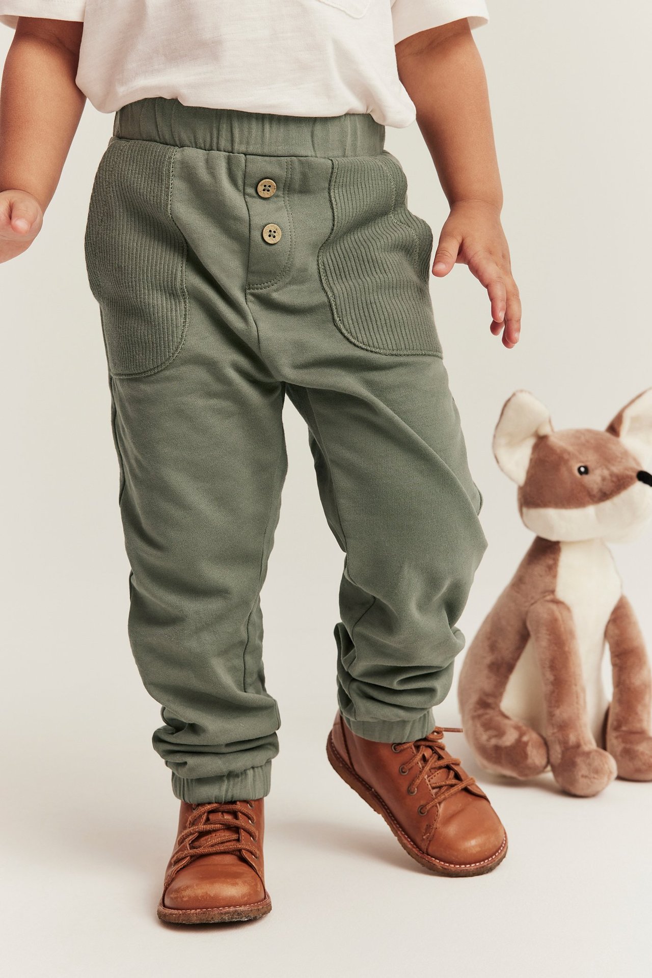 Vorderansicht: Baby in dunkelgrünen Baumwoll-Jogginghosen mit Ripptaschen, weißem T-Shirt und braunen Schuhen.