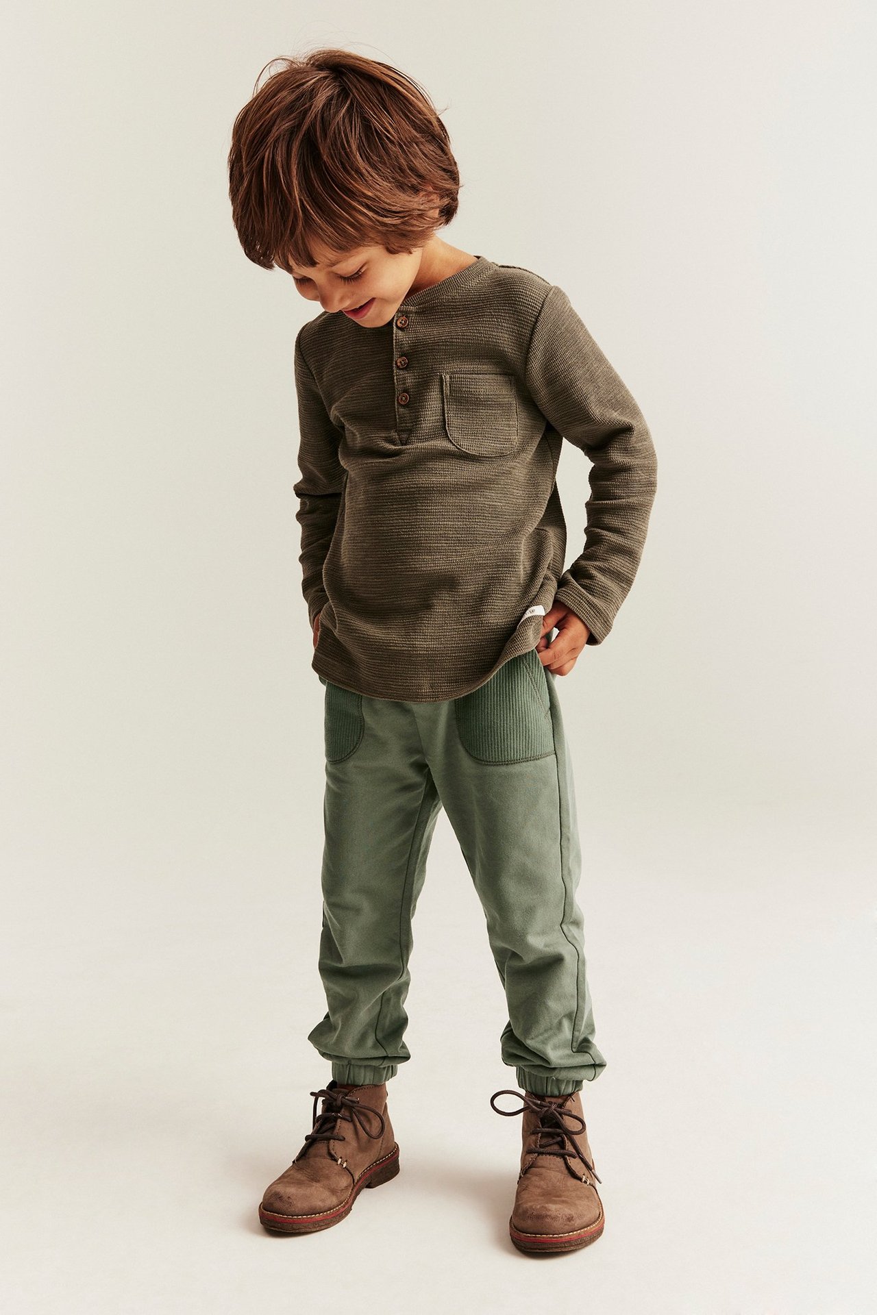 Vorderansicht: Ein Kind trägt dunkelgrüne Jogginghose mit Ripptaschen, ein dunkelolives Henley-Shirt und braune Stiefel.