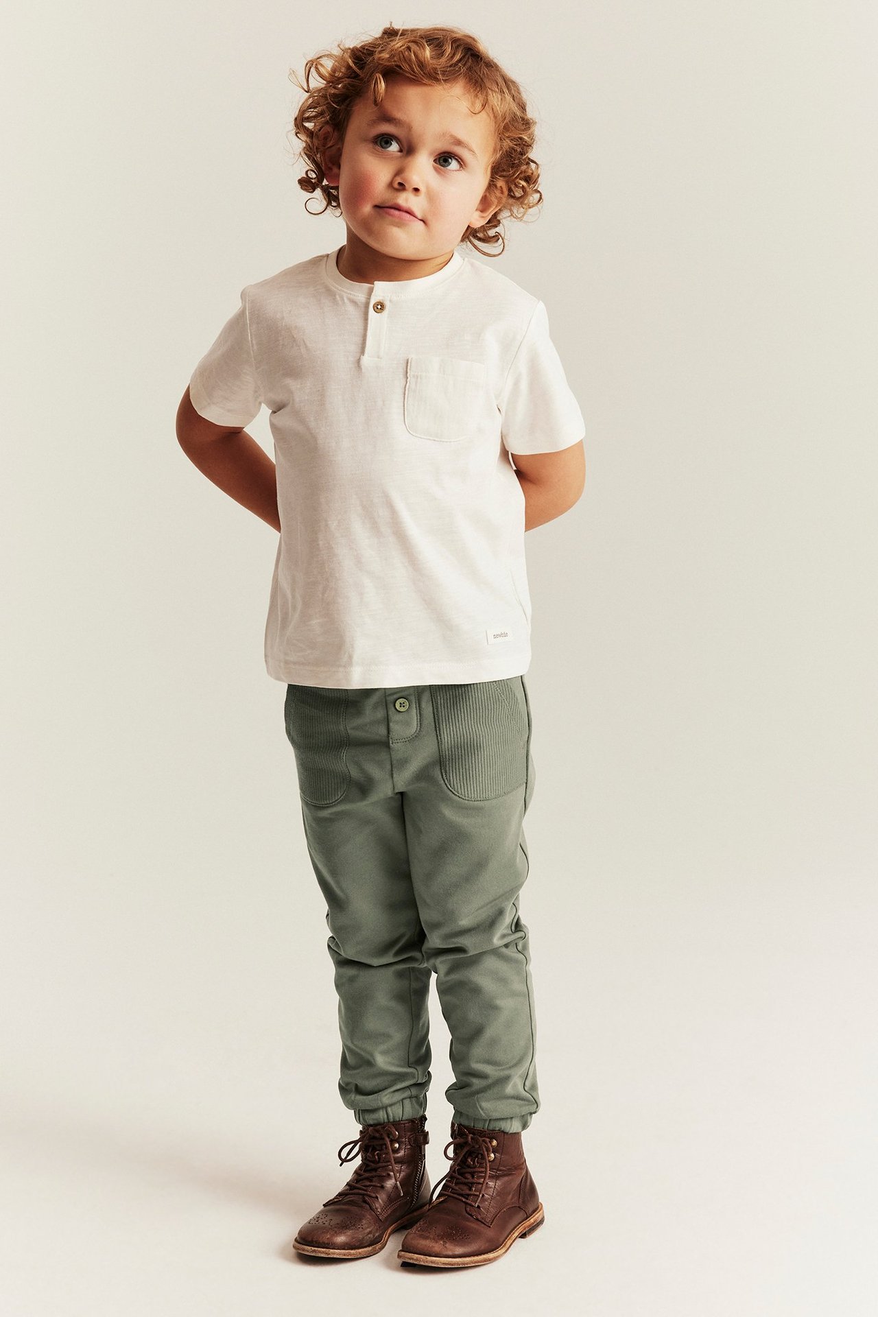 Front view of a kid wearing dark green cotton jogging pants, an off-white t-shirt, and brown boots.