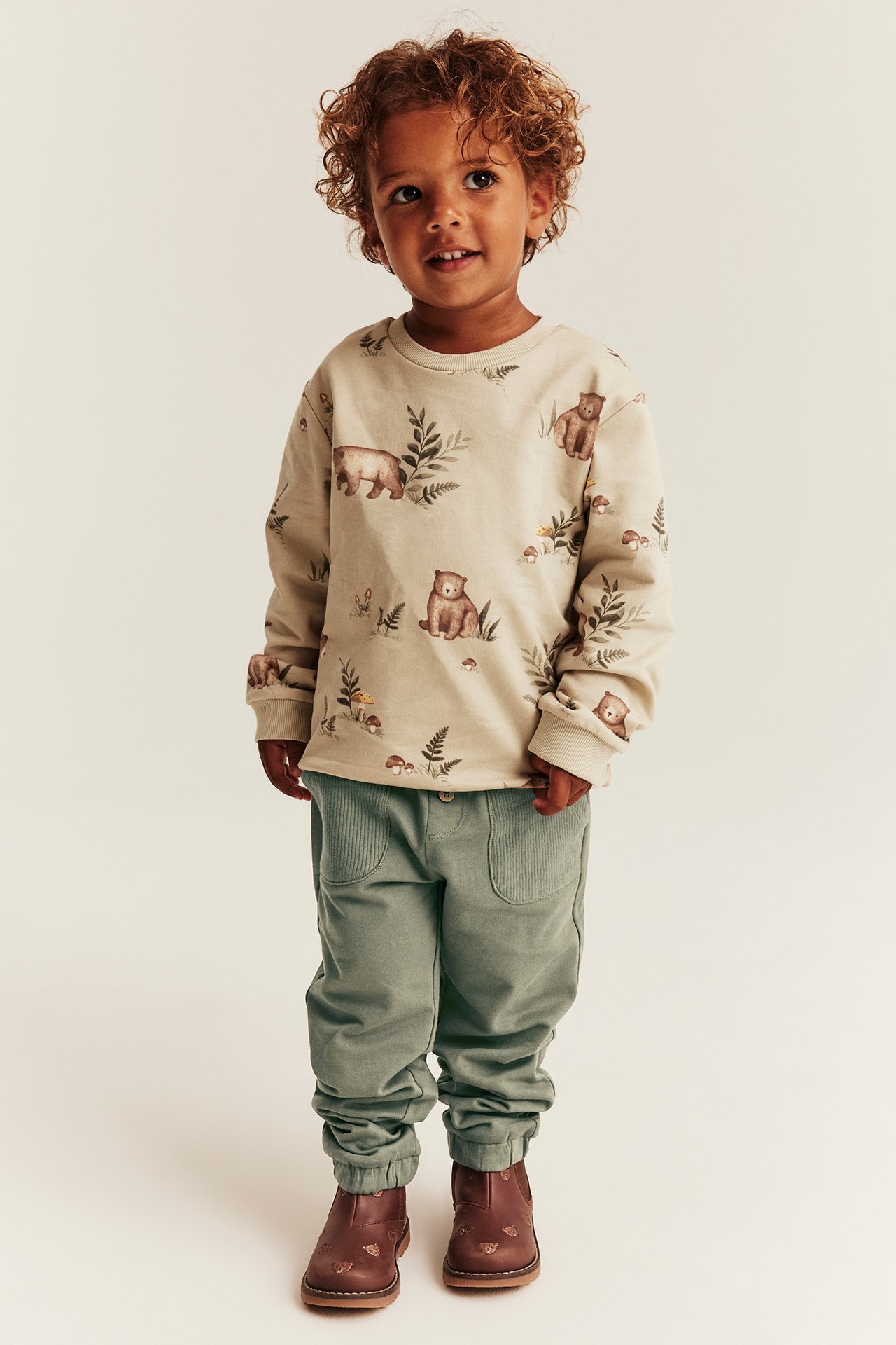 Front view: Kid in beige long-sleeve bear print top, sage green joggers, and brown boots.