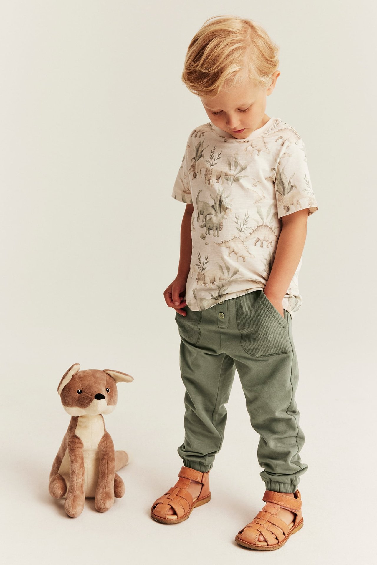 Front view: kid wears a cream dinosaur print t-shirt, green jogger pants, and brown sandals.