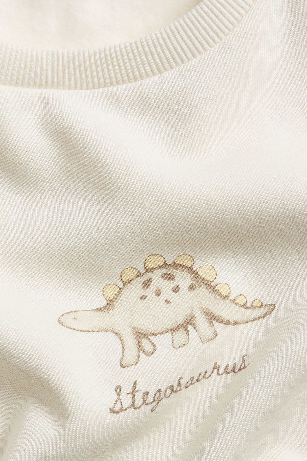 Nahaufnahme von einem cremefarbenen Baumwoll-Sweatshirt mit hellbraunem und gelbem Stegosaurus-Druck.