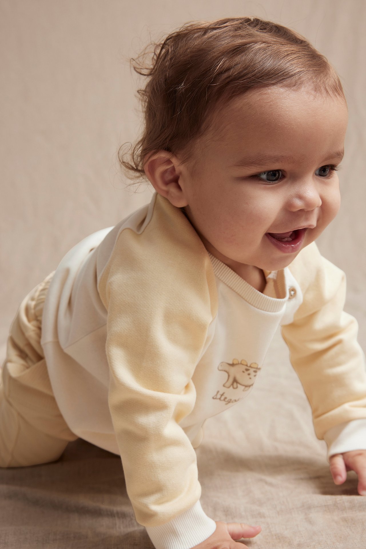 Front-side view of a baby in an off-white and light yellow dinosaur sweatshirt and light yellow pants.