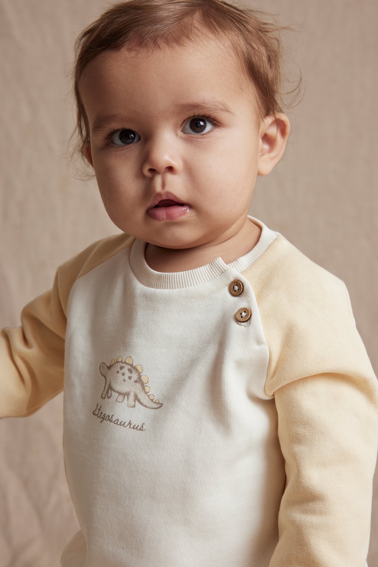 Close-up of a baby in an off-white and yellow cotton sweatshirt with dinosaur print and two buttons.