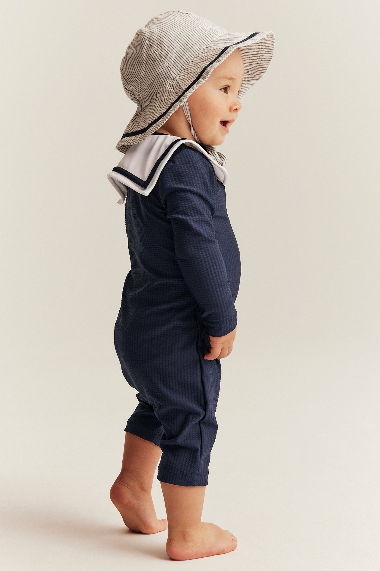 Seitenansicht eines Babys in dunkelblauem strukturiertem UV-Anzug mit weißem Matrosenkragen und gestreiftem Sonnenhut.