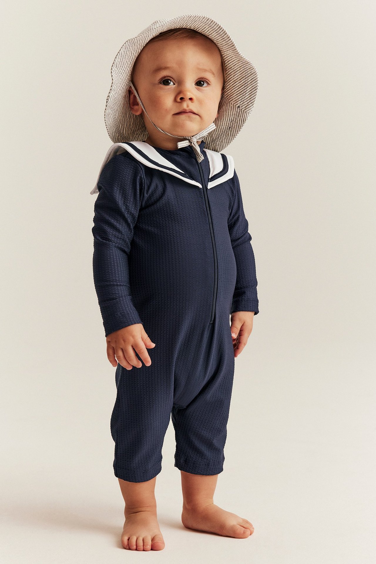 Vorderansicht Baby in dunkelblauem UV-Anzug mit weißem Matrosenkragen und gestreiftem Sonnenhut.