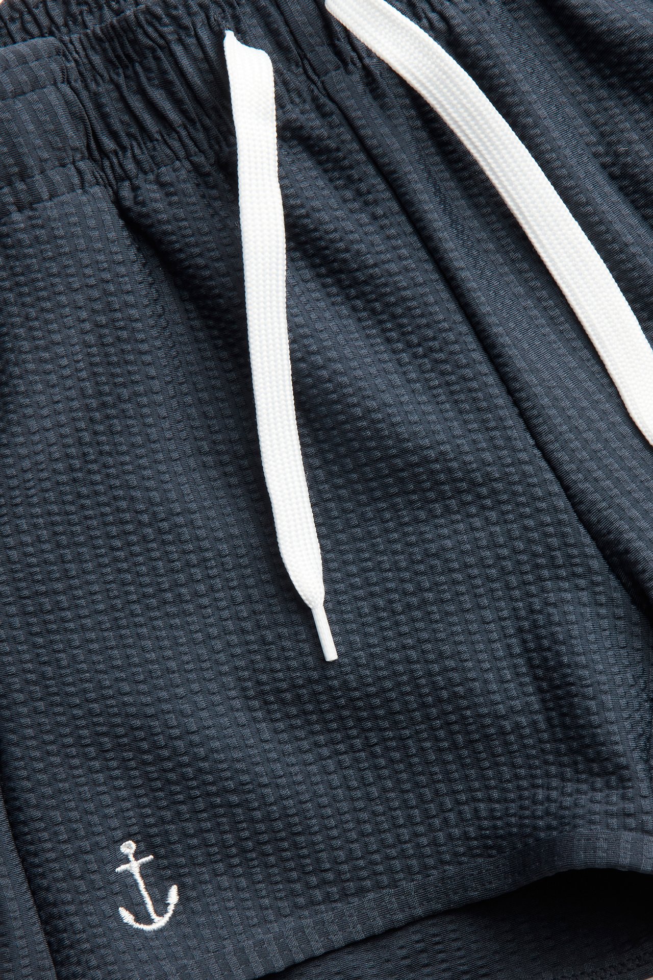 Nahaufnahme von dunkelblauen strukturierten Badeshorts mit weißem Kordelzug und Ankerstickerei.