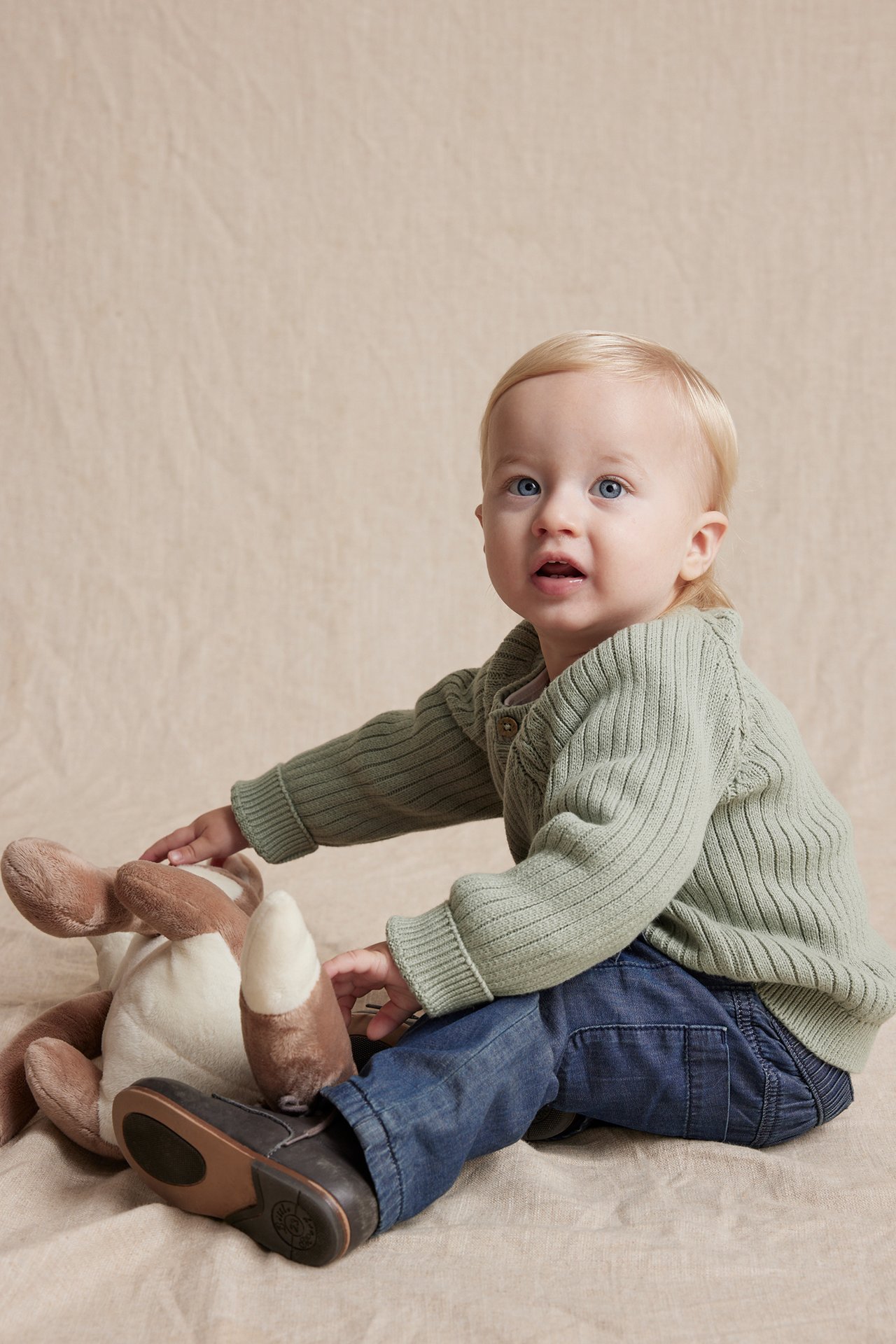 Baby in grünem Strickcardigan, blauen Jeans und dunklen Schuhen, spielt mit einem braunen Plüschtier.