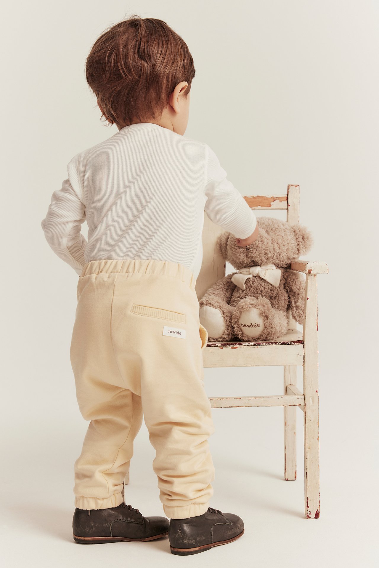 Rückenansicht eines Babys in hellgelben Baumwoll-Jogginghosen, einem weißen Ripp-Oberteil und dunkelbraunen Schuhen.