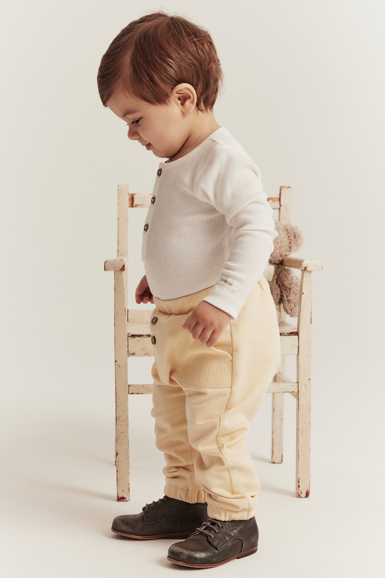 Seitenansicht eines Babys in gelben Jogginghosen mit Zierknöpfen, einem weißen gerippten Langarmshirt und dunkelbraunen Schuhen.