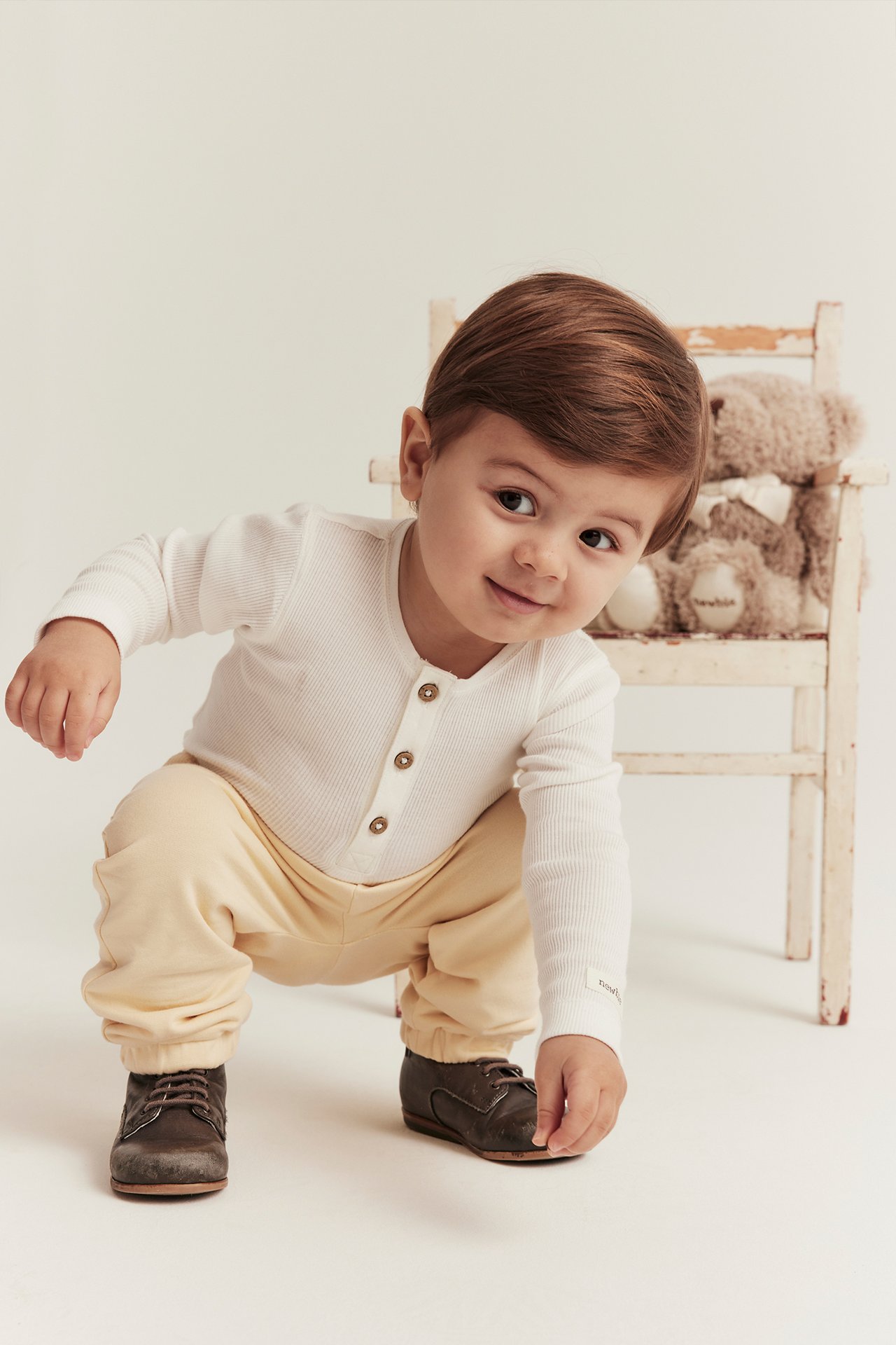 Vorderansicht eines Babys in gelben Baumwoll-Jogginghosen, einem weißen gerippten Langarmshirt und dunkelbraunen Schuhen.