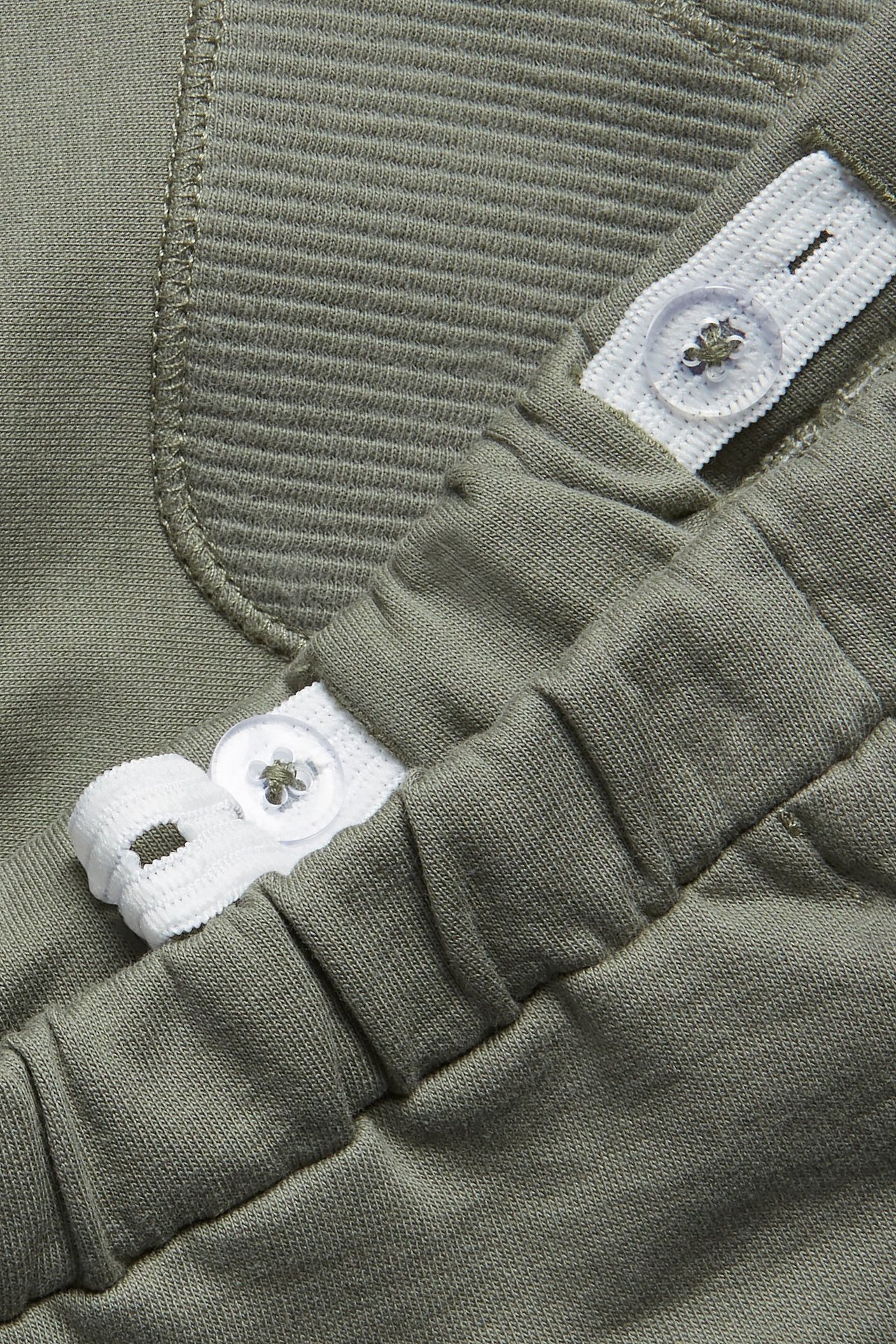 Close-up of dark green cotton jogging pants with ribbed detail, adjustable white elastic waist, and clear buttons.