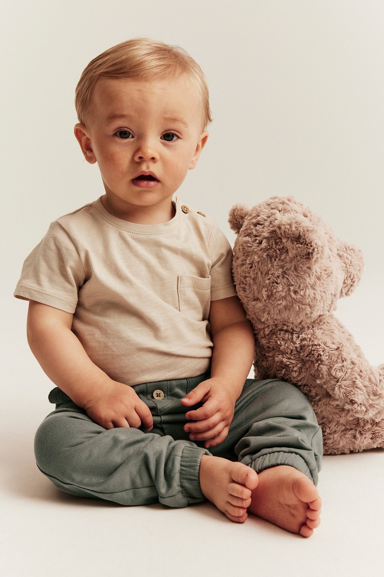 Front view of a baby wearing dark green cotton jogging pants, a beige t-shirt, and a brown teddy bear.