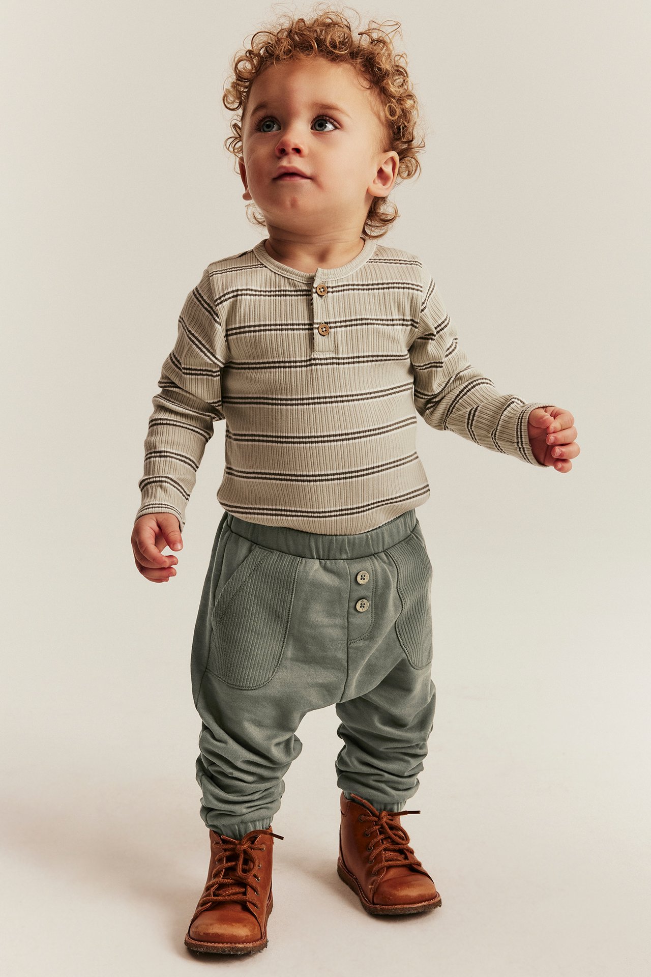 Vorderansicht: Baby in dunkelgrüner Jogginghose mit Knöpfen, gestreiftem Oberteil und braunen Stiefeln.
