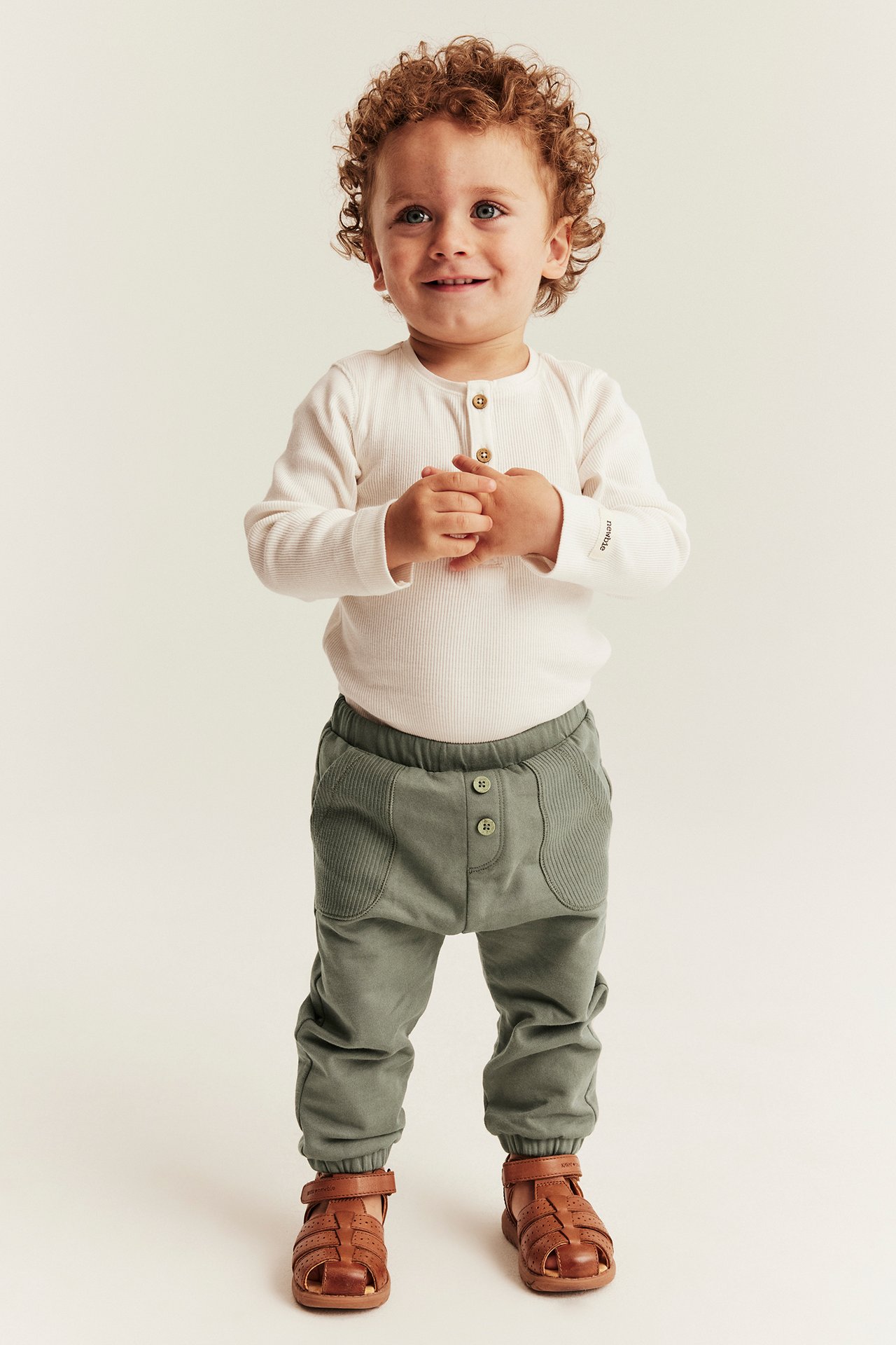 Front view of a kid wearing green jogger pants, a white ribbed henley shirt, and brown sandals.