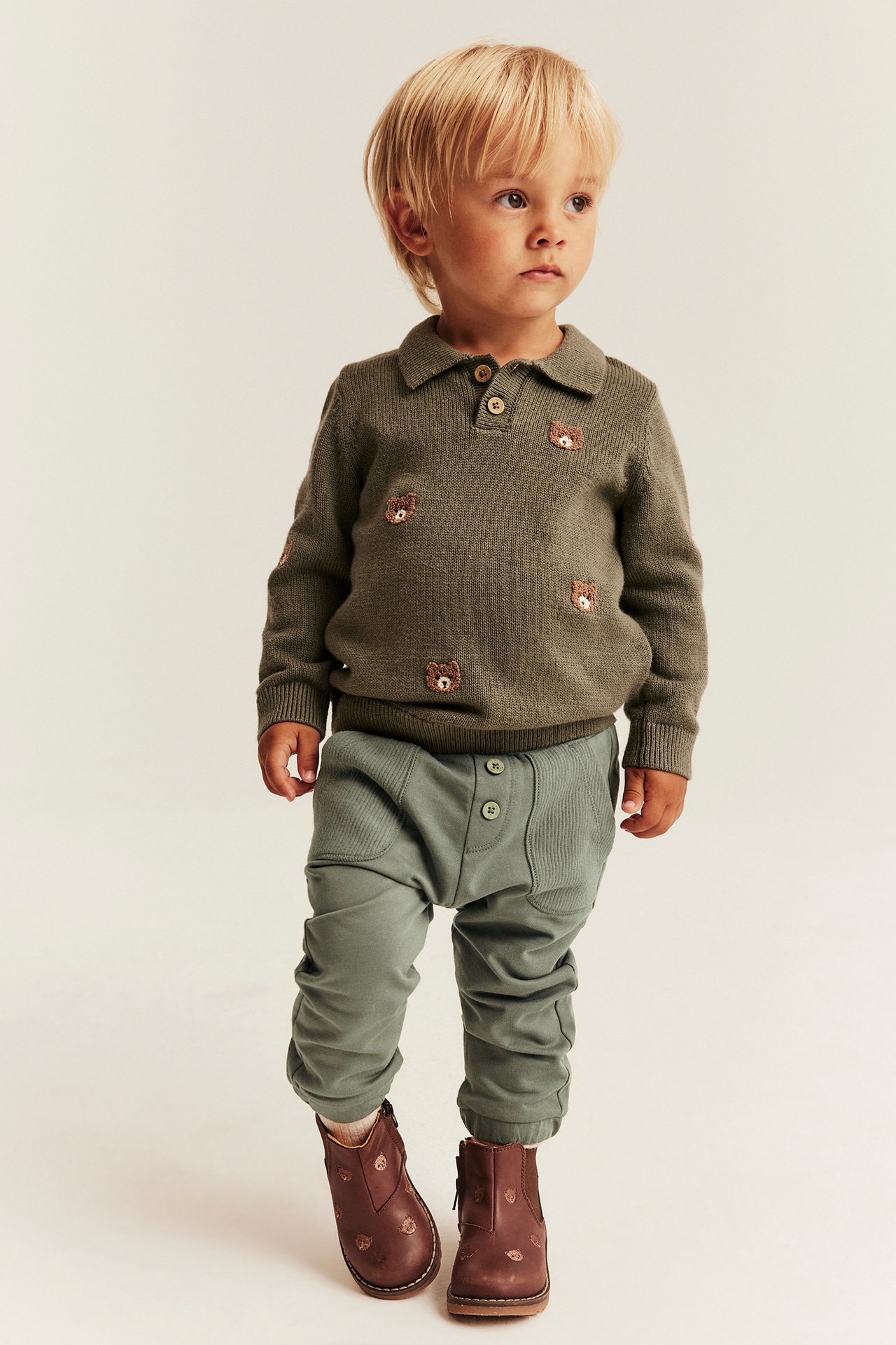 Kid in olive green knit polo sweater with bear embroidery, green pants, and brown boots.