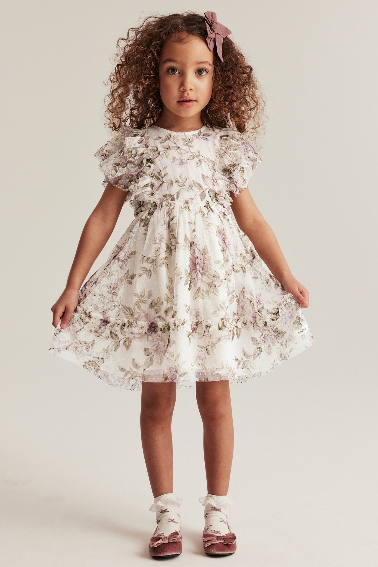Front view: Kid in off-white floral mesh dress with ruffled sleeves, pink velvet shoes, and a pink bow.