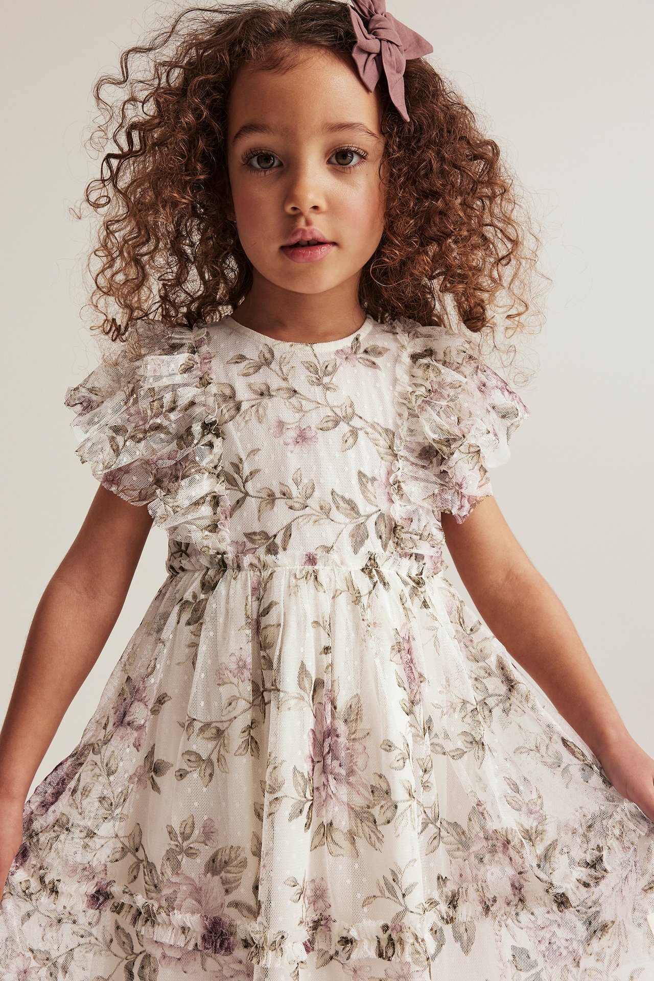 Front view of a kid in an off-white floral mesh dress with ruffled sleeves and a pink hair bow.